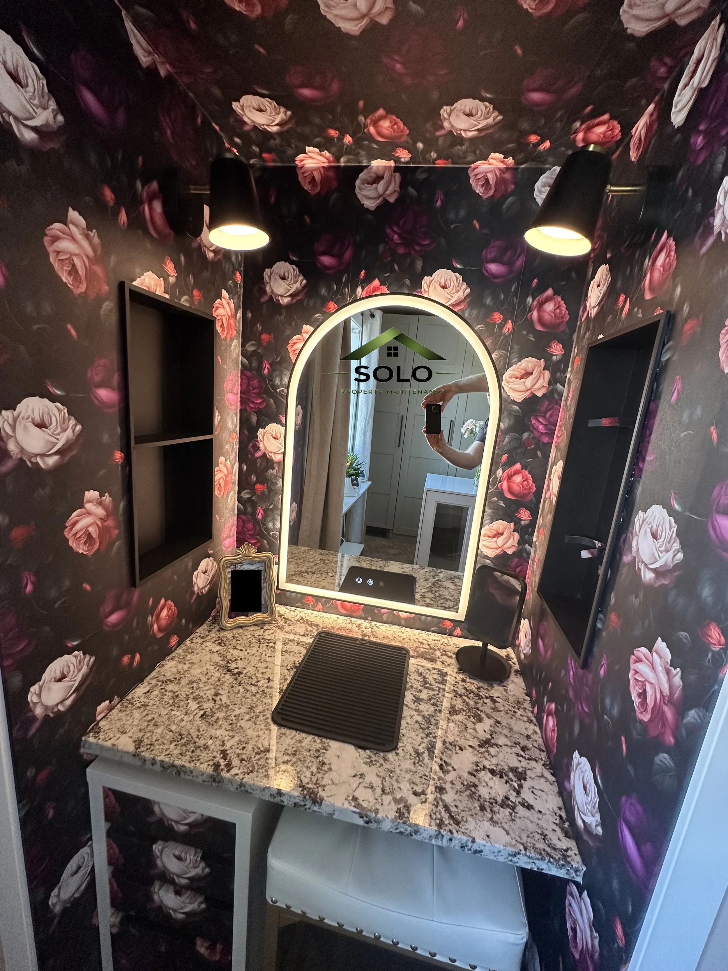 A bathroom vanity with a speckled granite countertop, a large backlit mirror, two built-in shelves on either side of the mirror, and a white cushioned stool underneath. There are black lamps directed at the mirror, and the wall behind has a dark flor