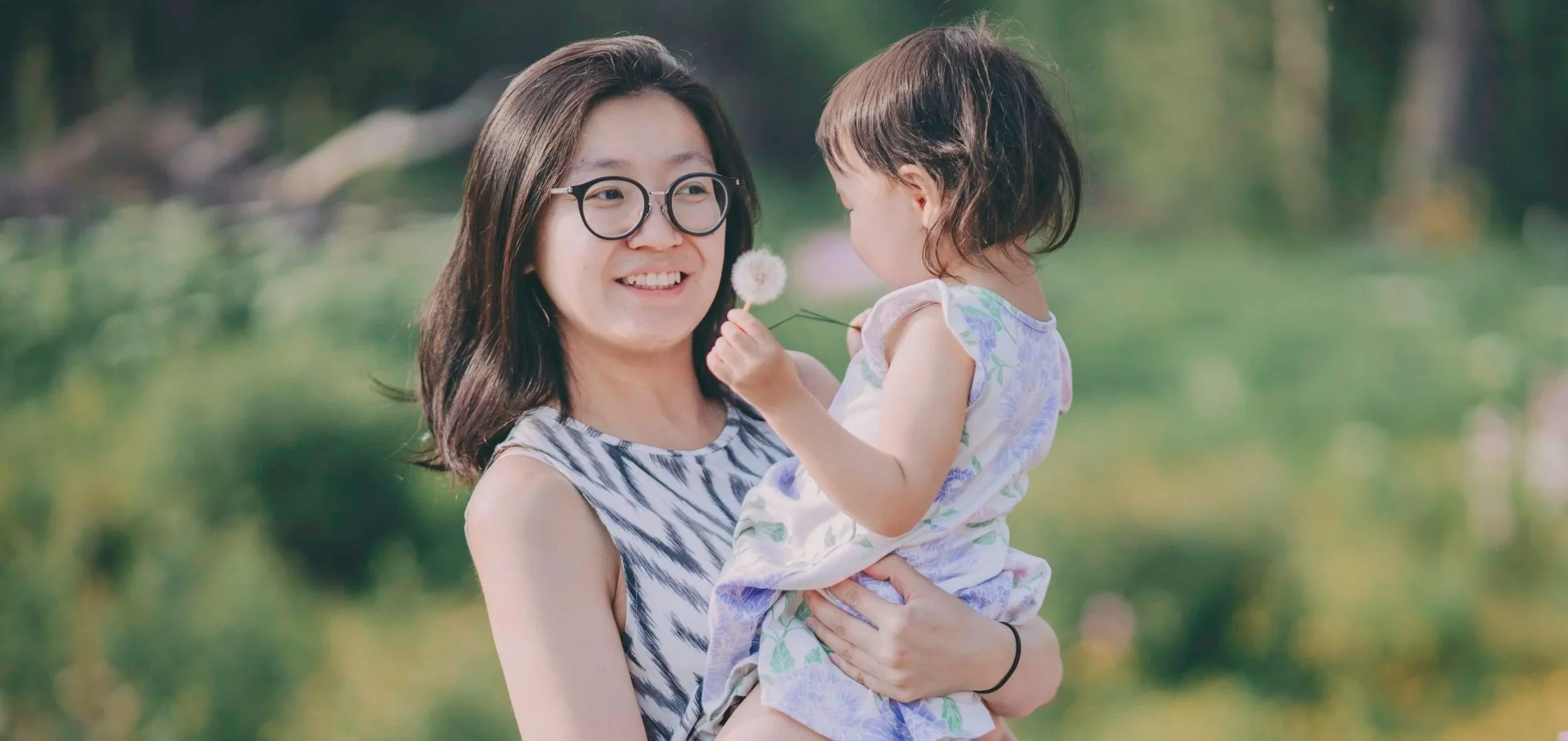 A woman with glasses holding a young girl in a park, the girl is holding a dandelion, and they are smiling at each other.