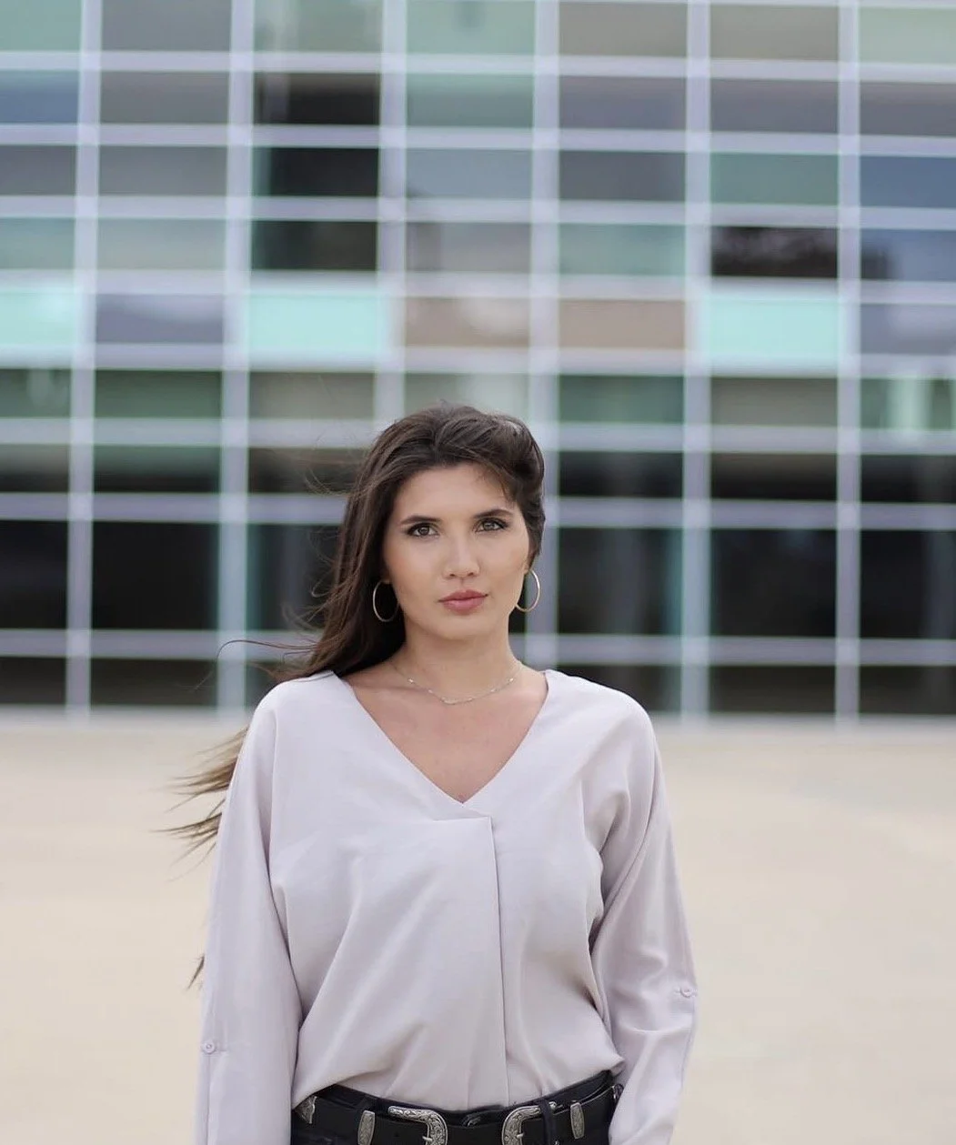 A woman with long dark hair, hoop earrings, and a light-colored blouse standing in front of a modern glass building.