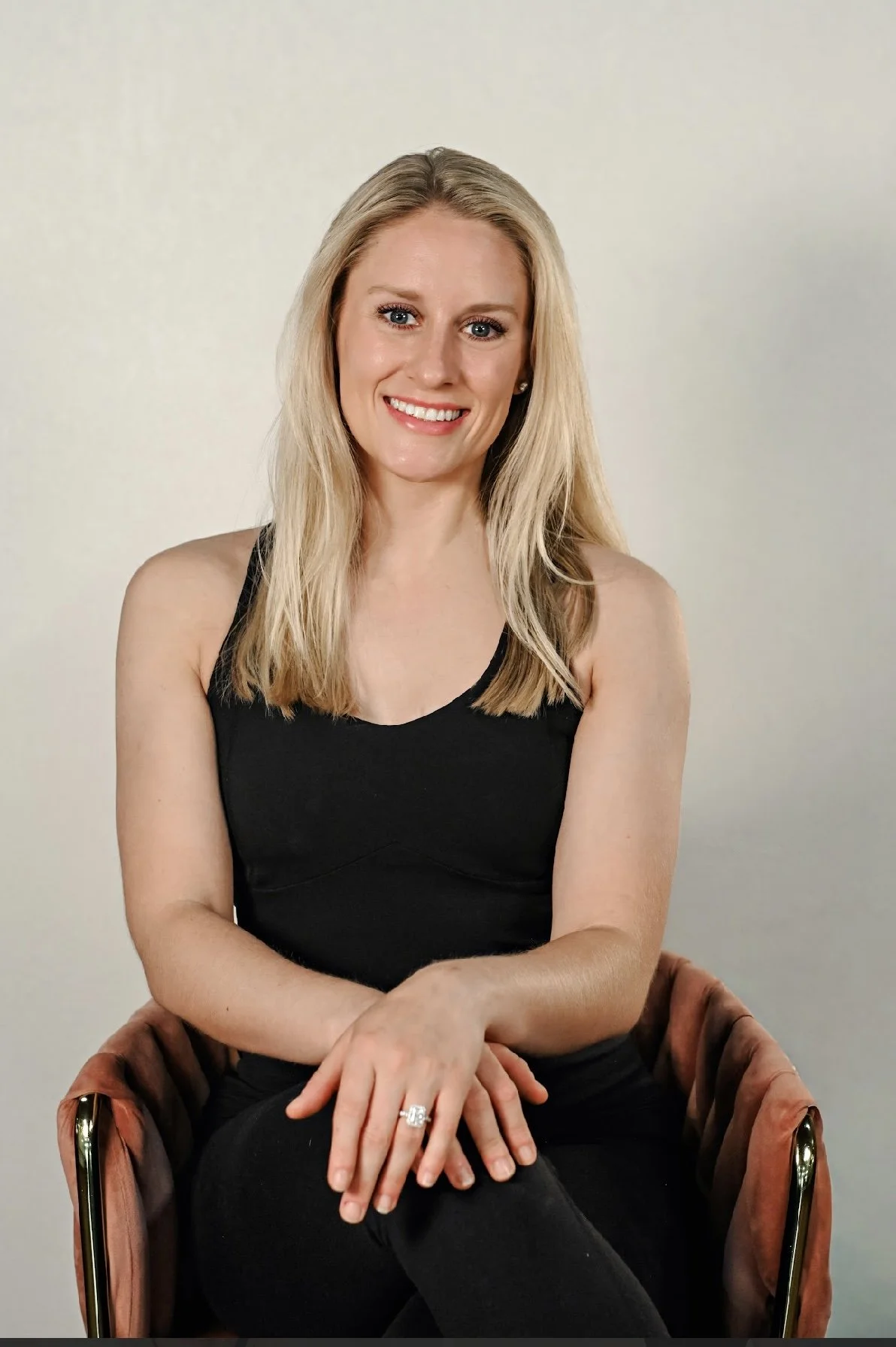 A smiling blonde woman wearing a black sleeveless top, sitting on a chair with her hands rested on her lap, against a plain off-white background.
