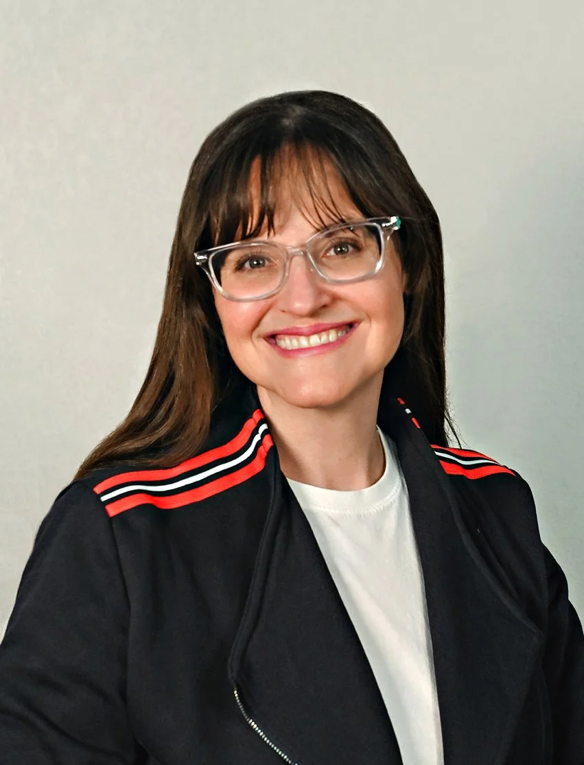 A woman with shoulder-length brown hair, glasses, and a friendly smile, wearing a black jacket with red and white stripes on the shoulders, over a white shirt, standing against a plain wall.