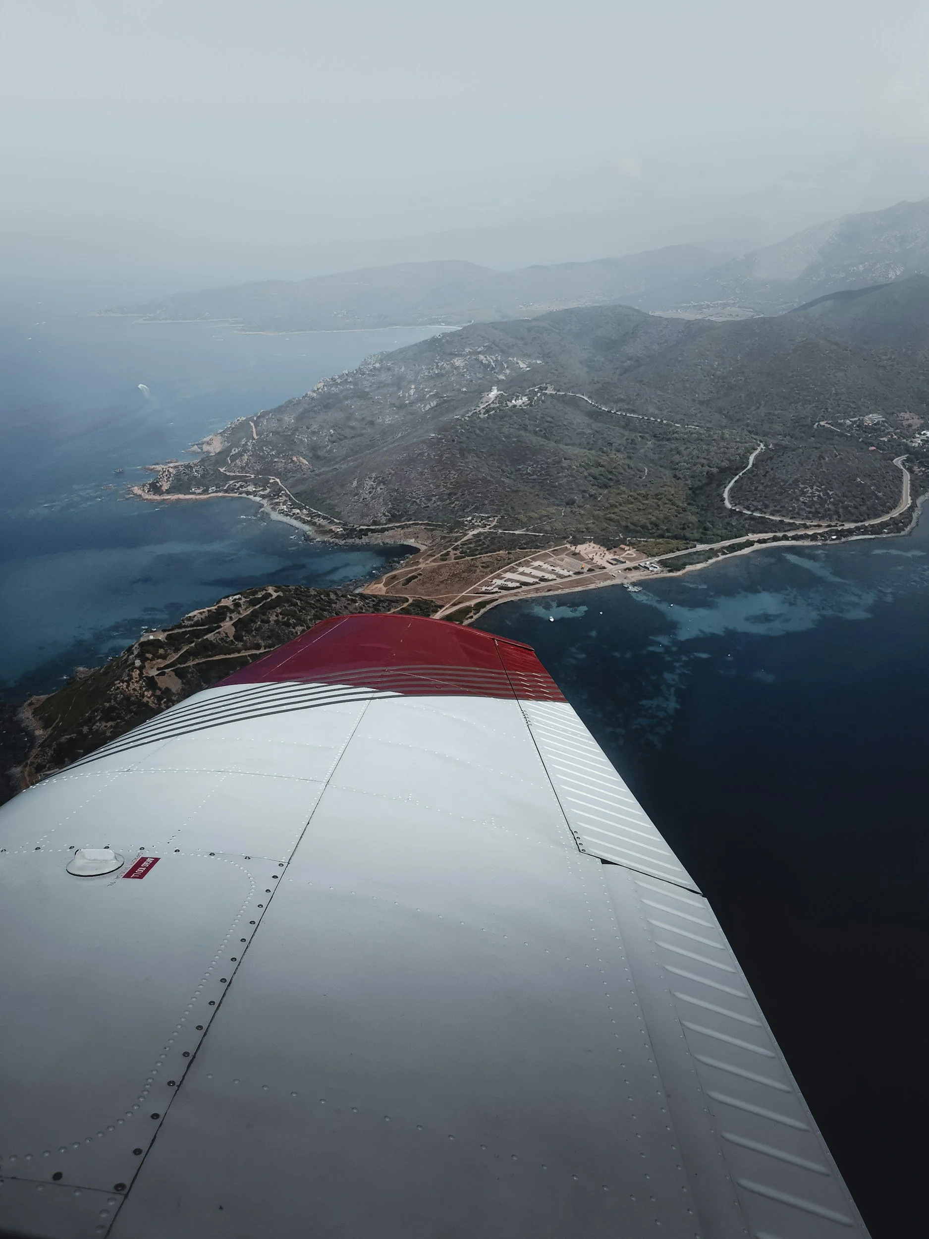 Aerial view of coastline with mountains, water, and a small scattered settlement, seen from a red and white aircraft wing.