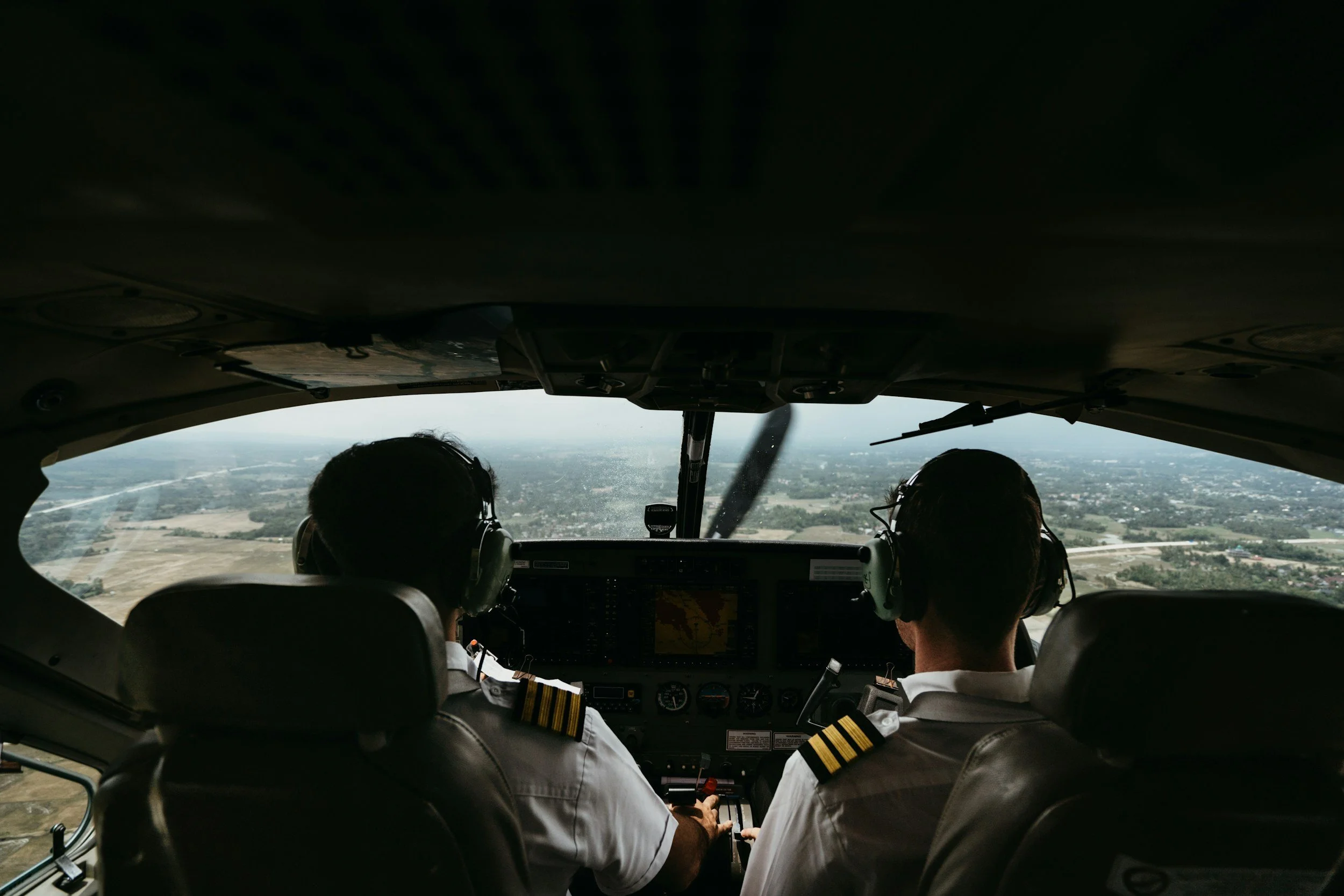 Two pilots sitting in the cockpit of a small airplane, flying over a landscape with fields and roads.