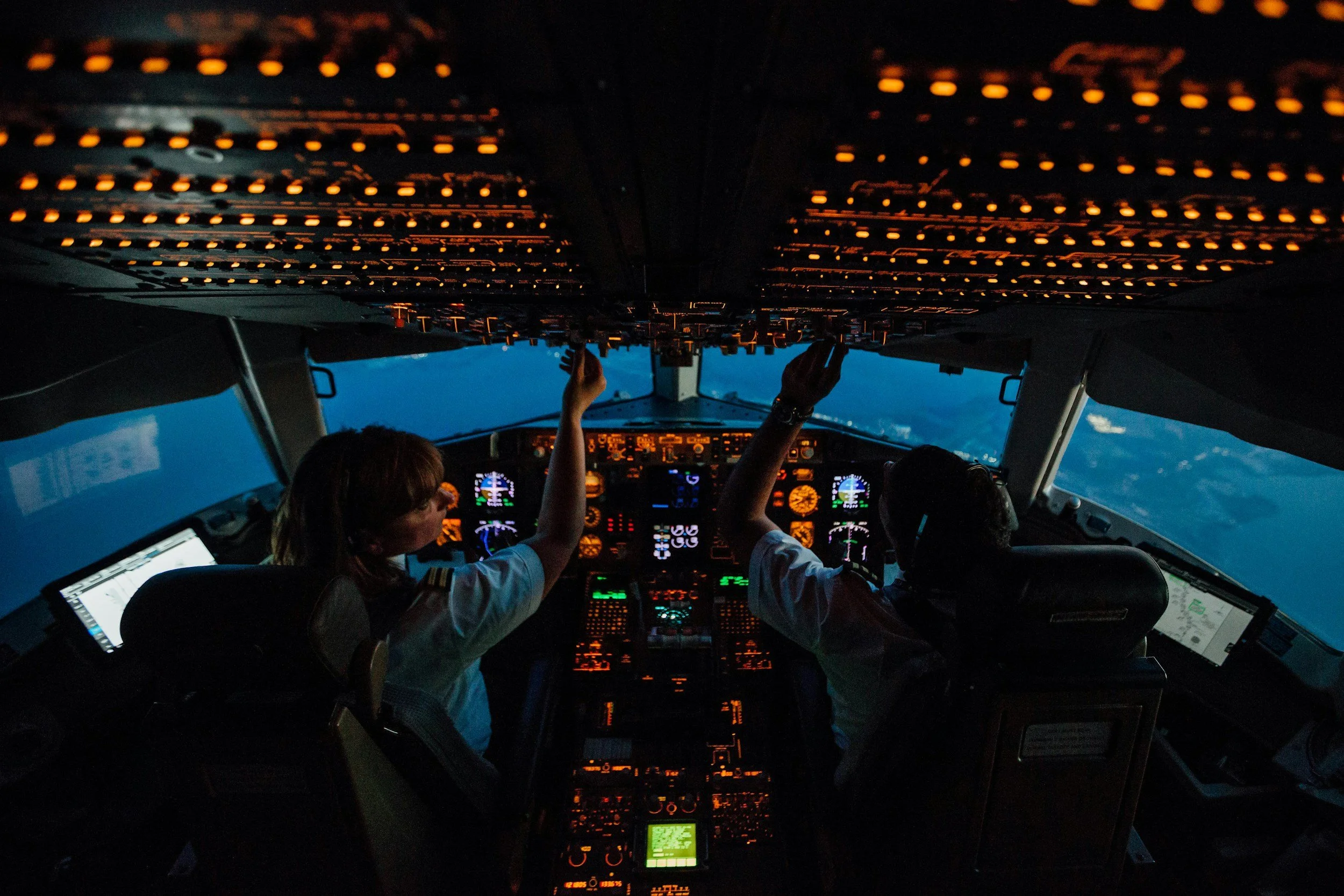 Inside the cockpit of an airplane, two pilots wearing white uniforms with shoulder patches are operating the controls during twilight or night. The cockpit is illuminated by numerous switches and gauges, with a view of the sky visible through the front windows.