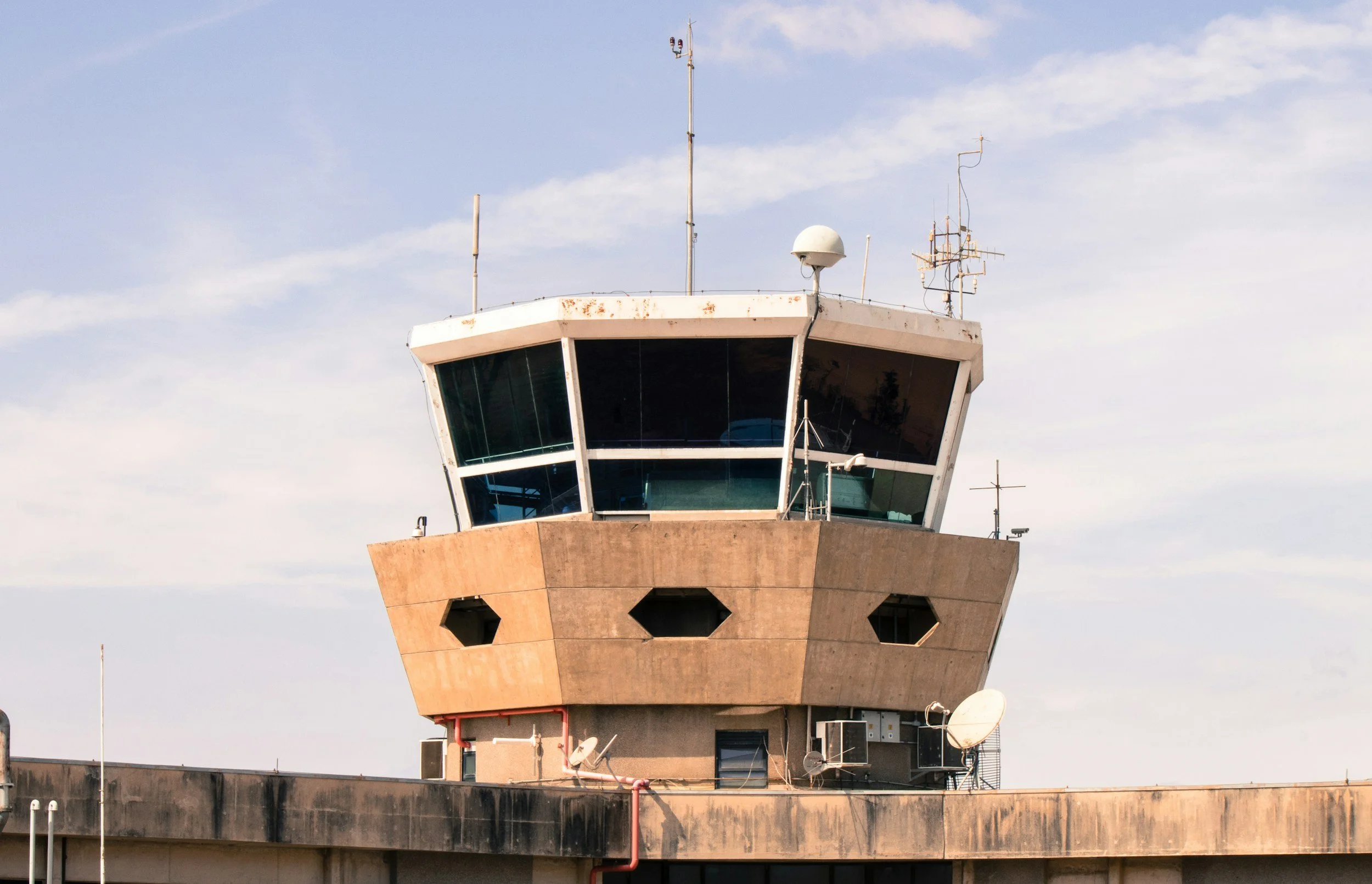 Close-up of an airport control tower with multiple antennas and satellite dishes on the roof, against a partly cloudy sky.