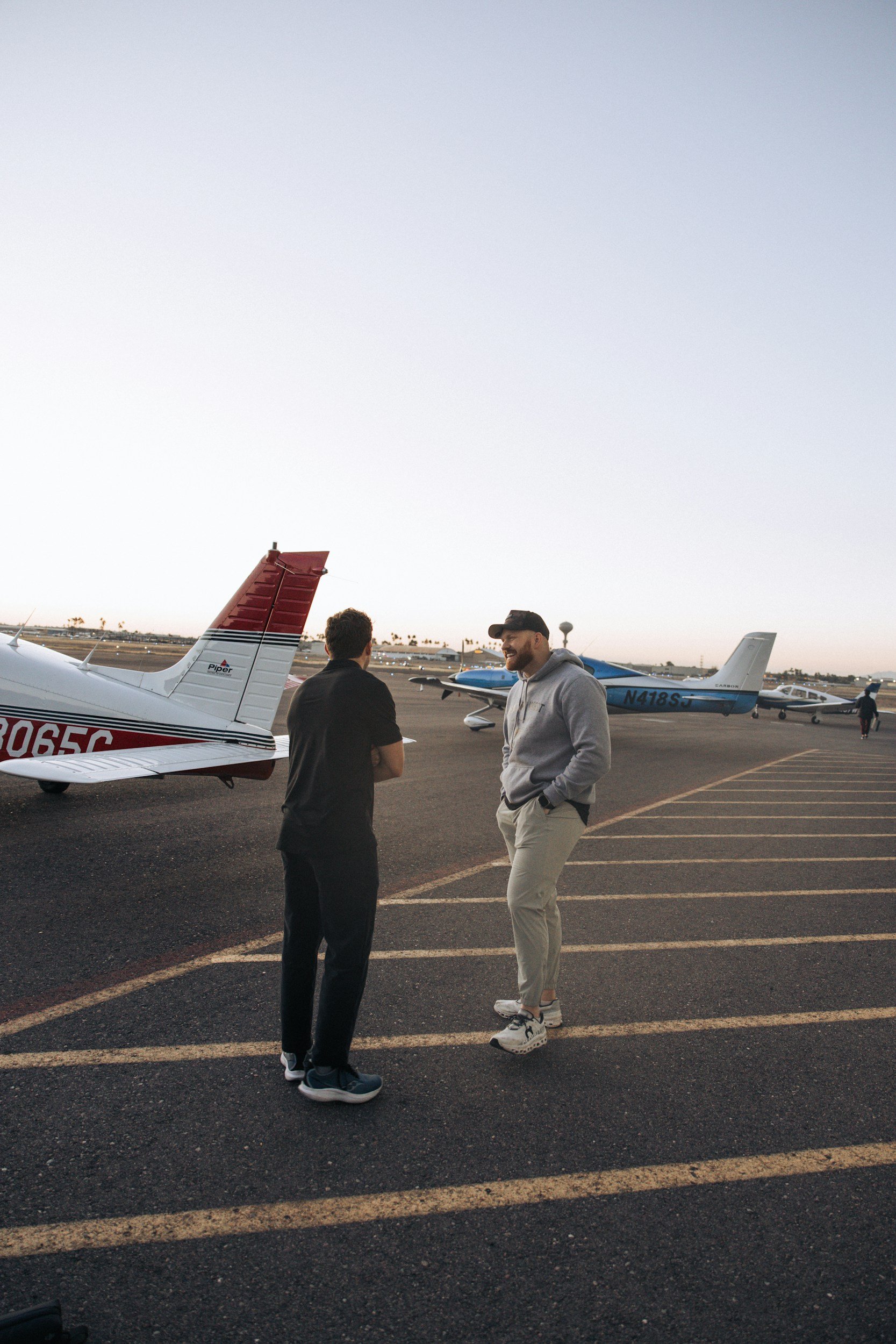 Two men standing on an airport tarmac near small airplanes, engaging in conversation with other planes and airport facilities visible in the background during dusk.
