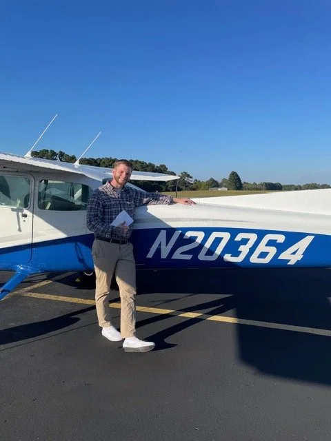 A man standing next to a small blue and white airplane with the tail number N20364 on a paved airstrip, holding a paper and pointing to the plane under a clear blue sky.