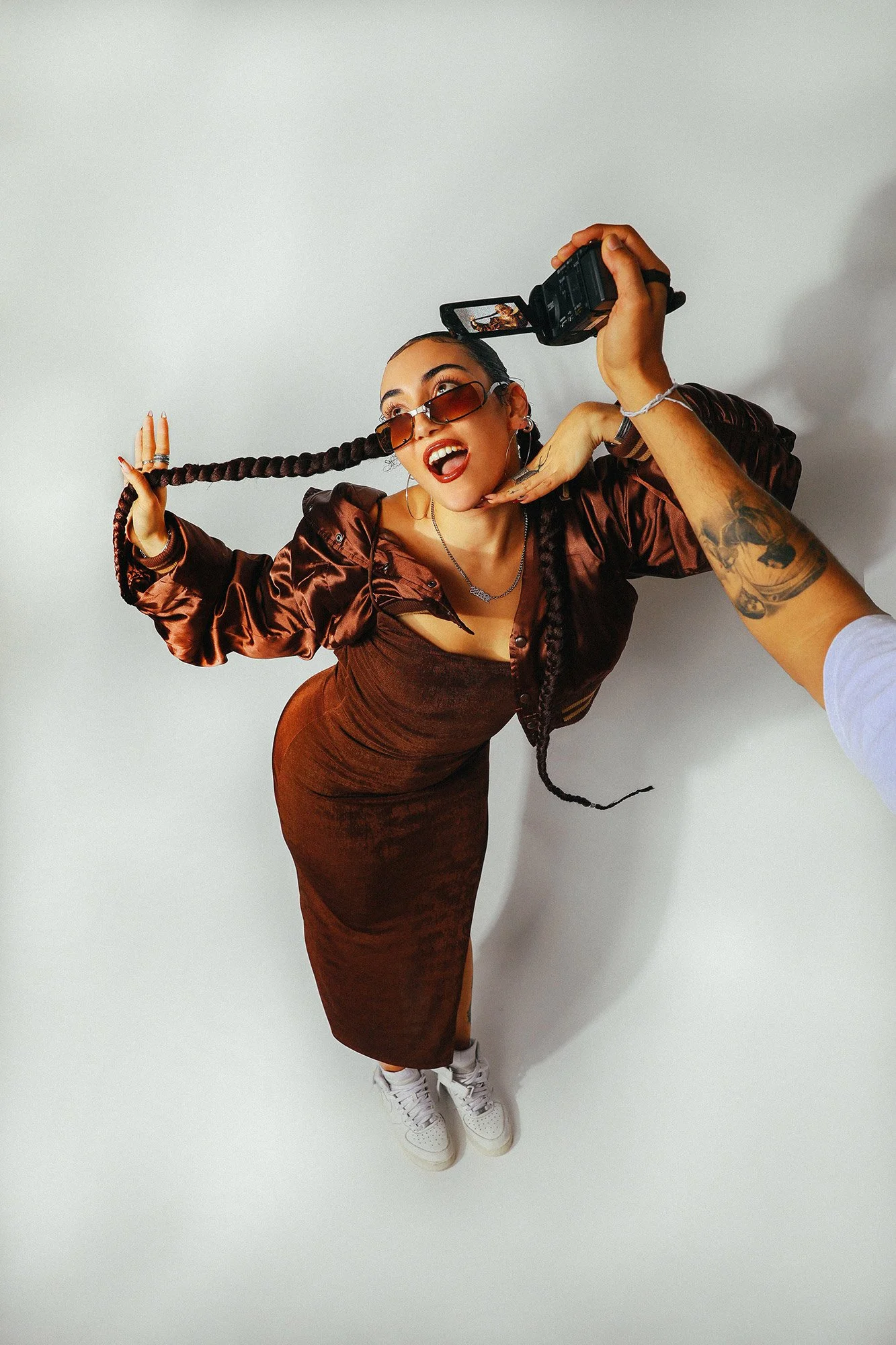 Une femme portant une robe marron, des lunettes de soleil, et des bijoux, pose en mode selfie contre un fond blanc, en souriant et avec une coiffure à longues tresses.