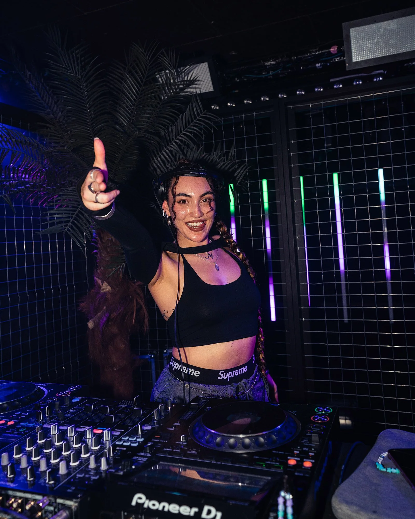 Une femme DJ souriante, portant un t-shirt noir et un pantalon à motif, joue avec ses platines dans une ambiance de fête avec des lumières colorées et des décorations tropicales en arrière-plan.