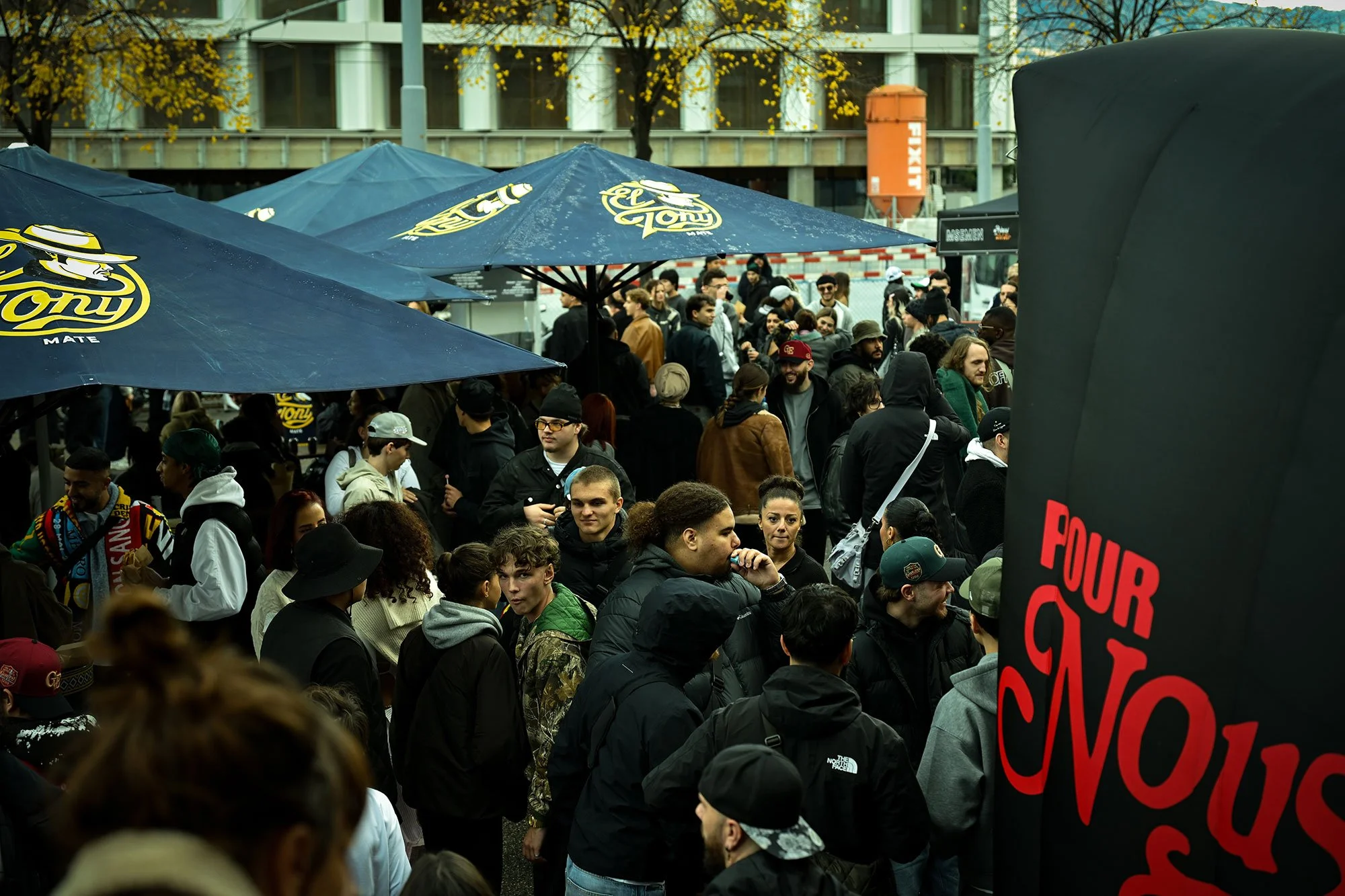 Une foule de personnes rassemblées dans une rue sous des parapluies bleus portant le logo de la marque de bière Tony. Les gens semblent être en attente ou en train de participer à un événement public, avec des arbres en arrière-plan et un bâtiment moderne.