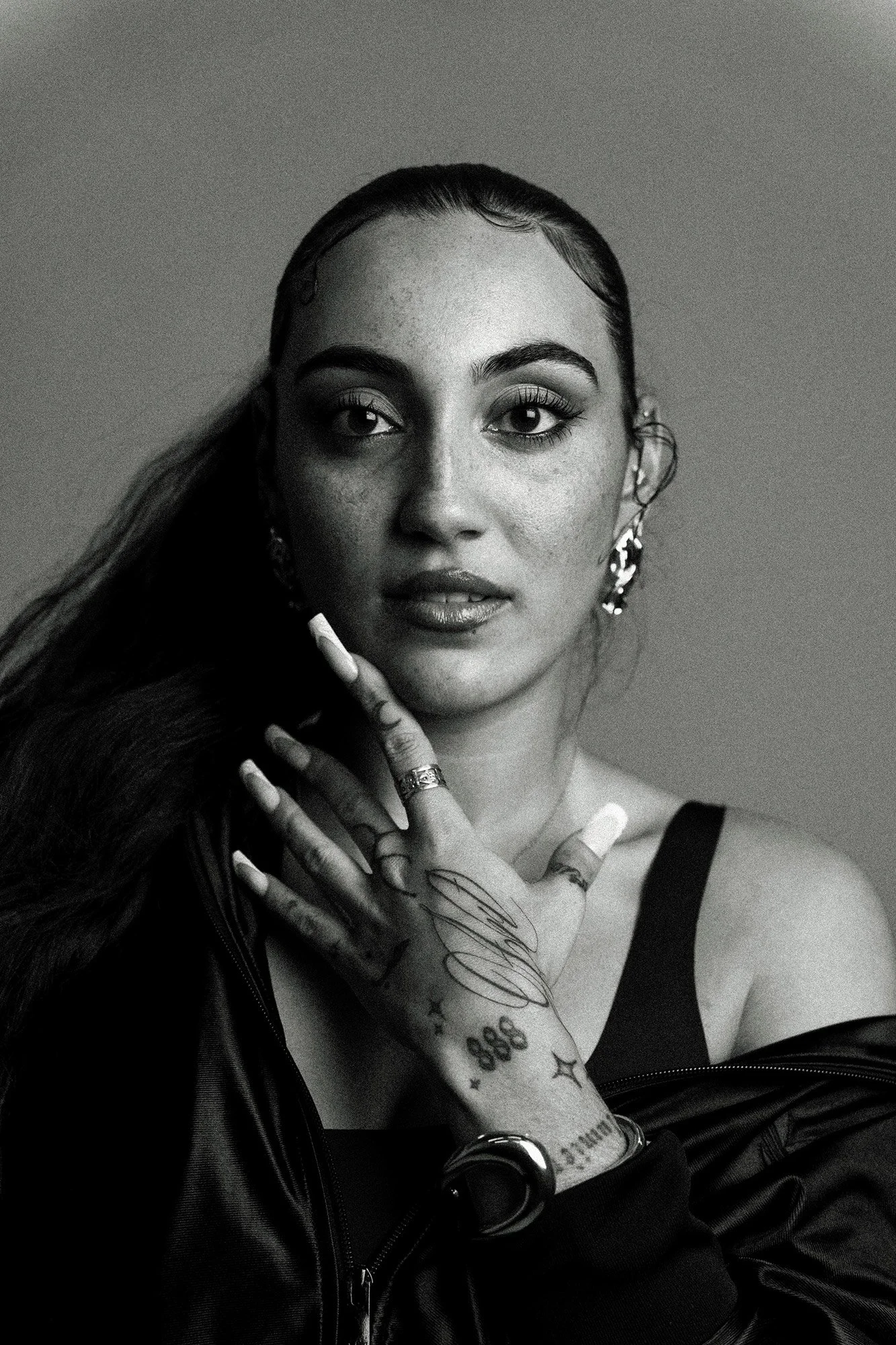 Portrait en noir et blanc d'une femme avec de longs cheveux, portant des boucles d'oreilles, des tatouages sur la main et le bras, et un haut noir, posant avec une expression sérieuse.