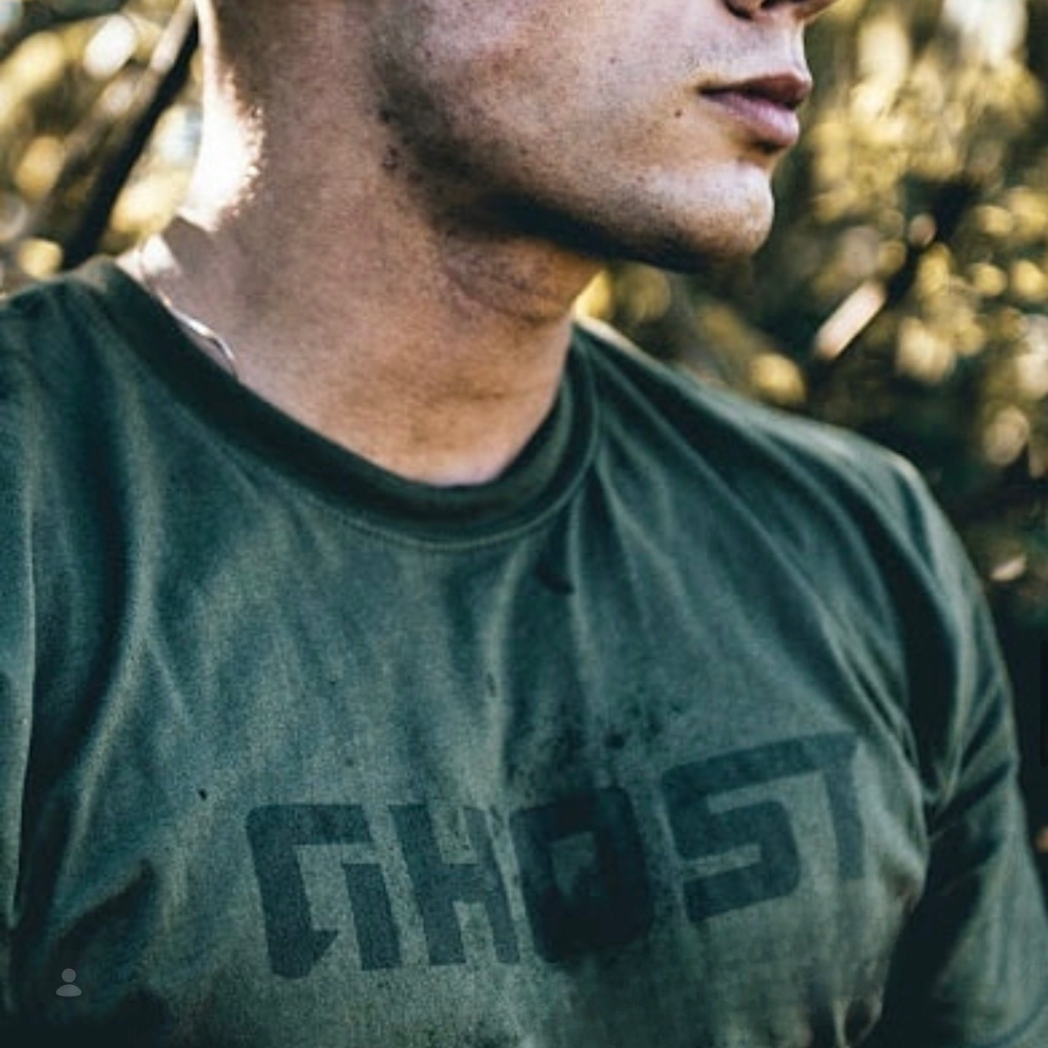 Close-up of a person's face and upper body, wearing a dark green T-shirt outdoors with blurred trees in the background.