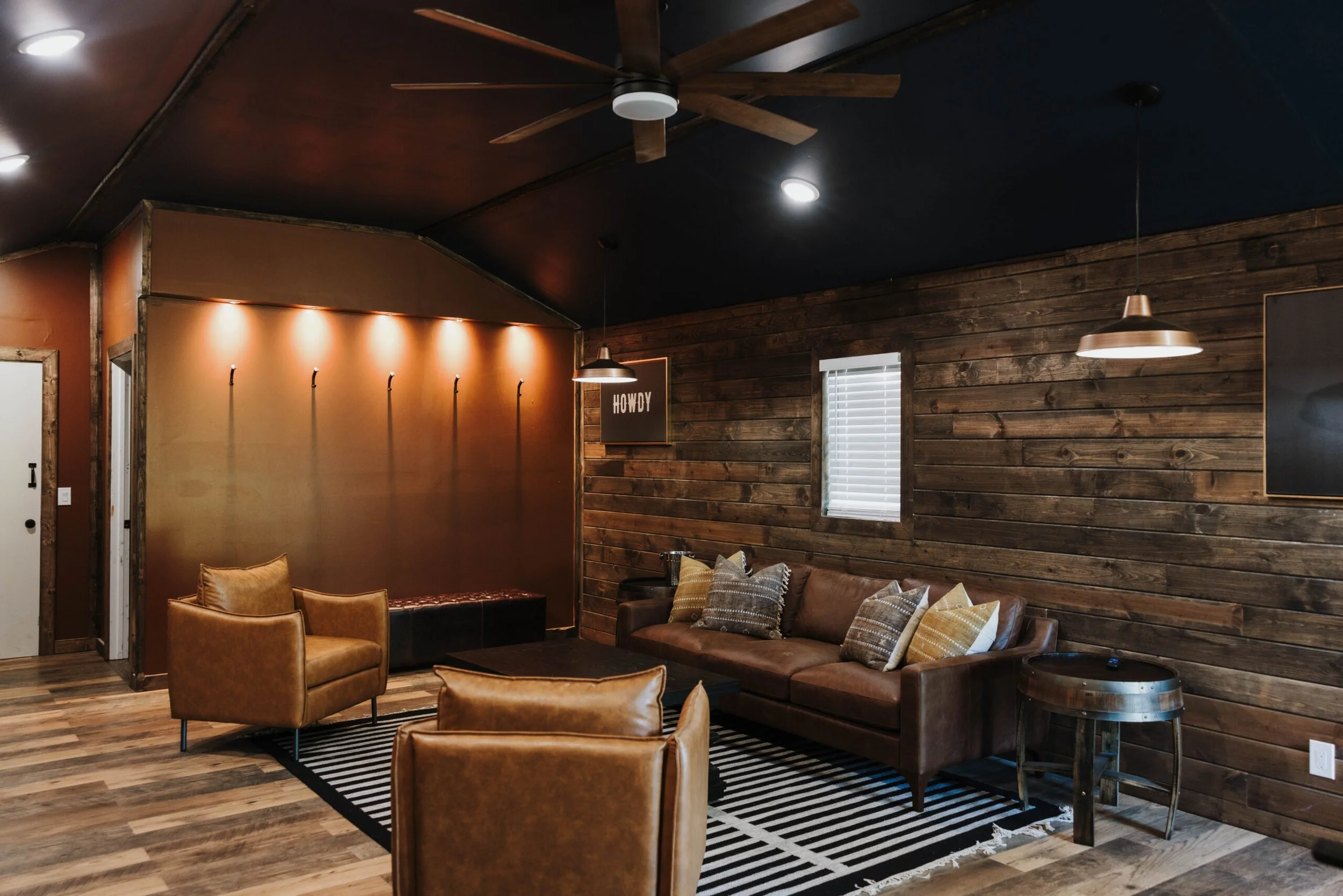 A cozy lounge area with wooden walls, a leather sofa with pillows, a striped rug, and a corner with chairs. The ceiling has a modern fan and recessed lighting, and there are windows with blinds assessing natural light.