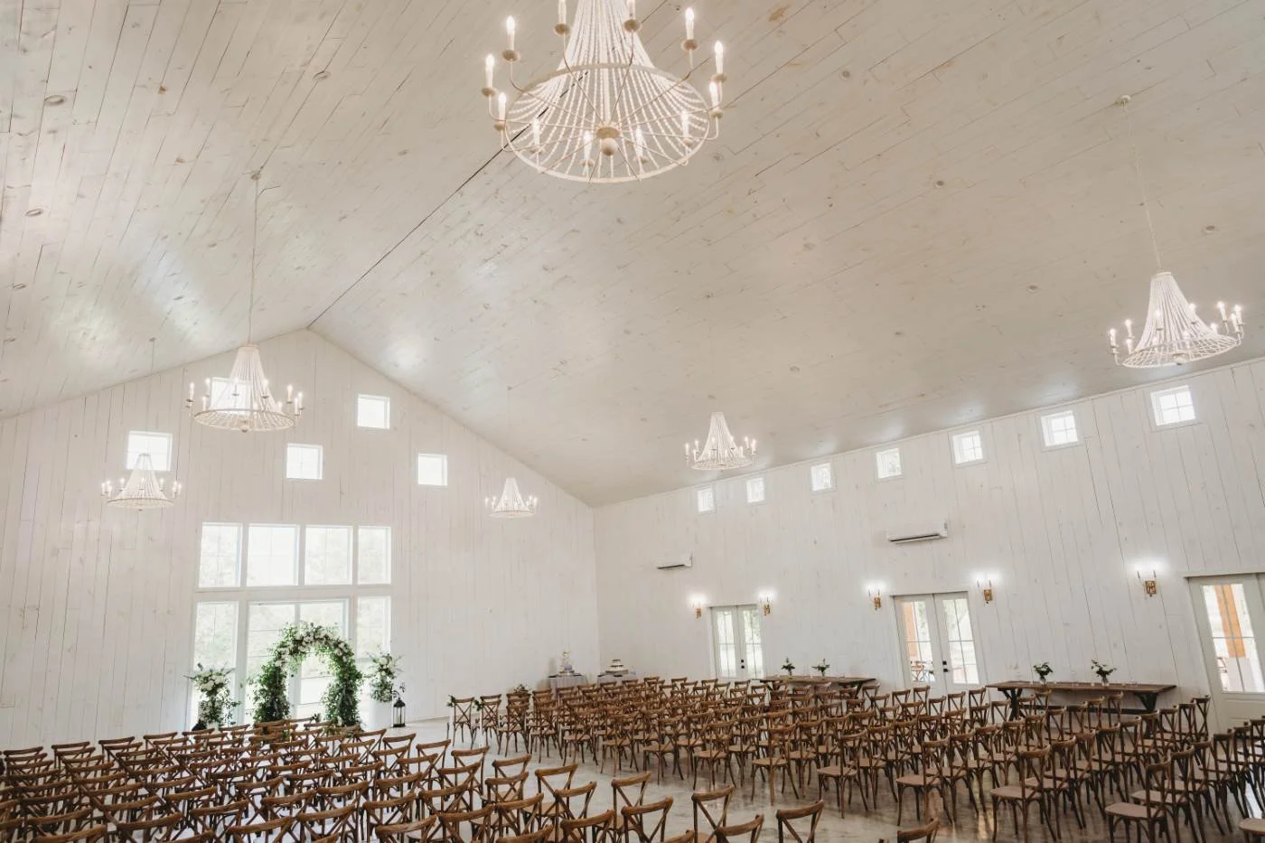 A spacious wedding ceremony hall with white wooden walls and ceiling, rows of wooden chairs, large window with floral arch, chandeliers hanging from ceiling, and side doors leading outside.