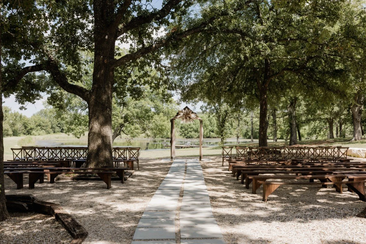 Outdoor wedding setup with a white aisle runner, wooden benches on either side, a wooden arch decorated with flowers under large trees, and a scenic lake in the background.