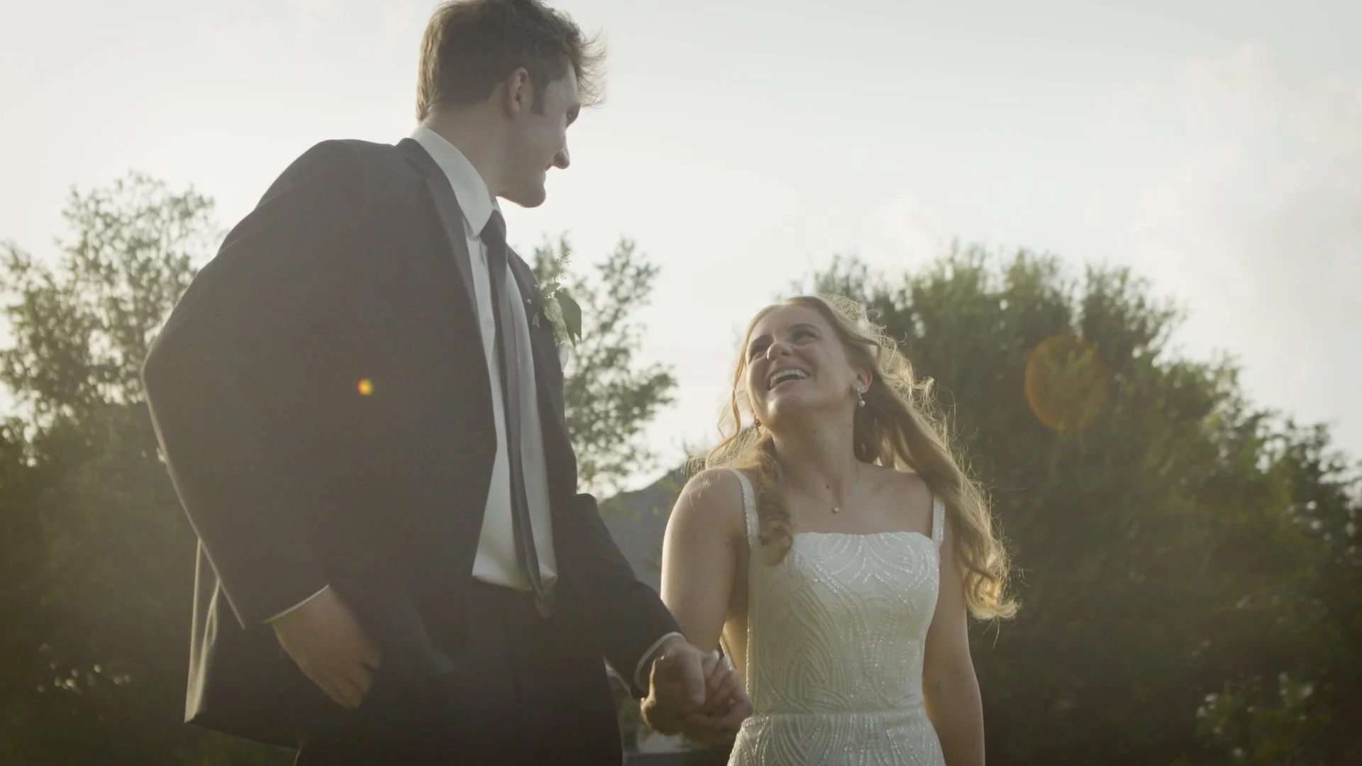 A wedding couple holding hands outdoors, smiling at each other, with trees and sunlight in the background.