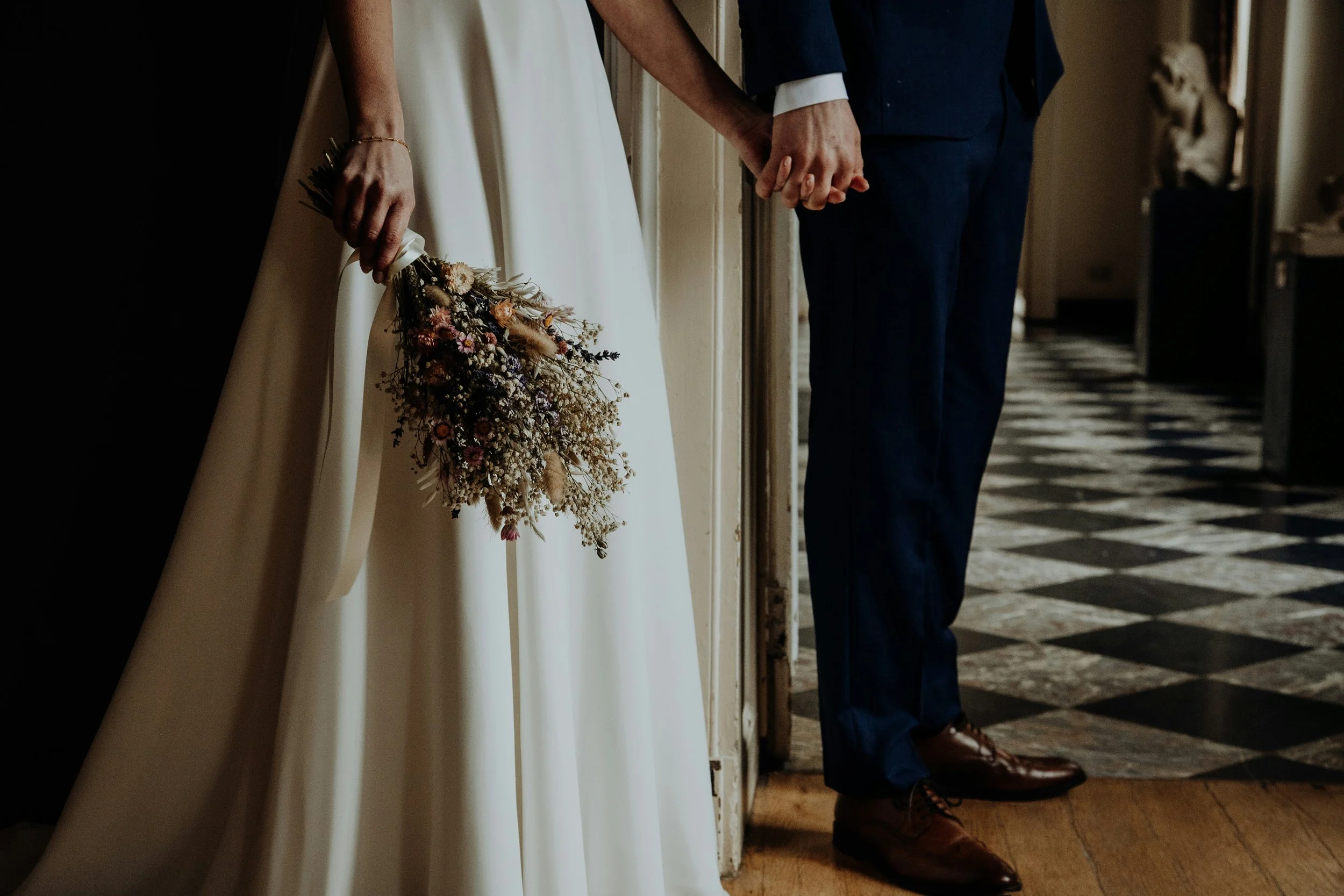 Close-up of a couple holding hands, standing side by side, with the woman holding a bouquet of flowers and the man wearing a dark suit with brown shoes, in a dimly lit hallway with patterned flooring.
