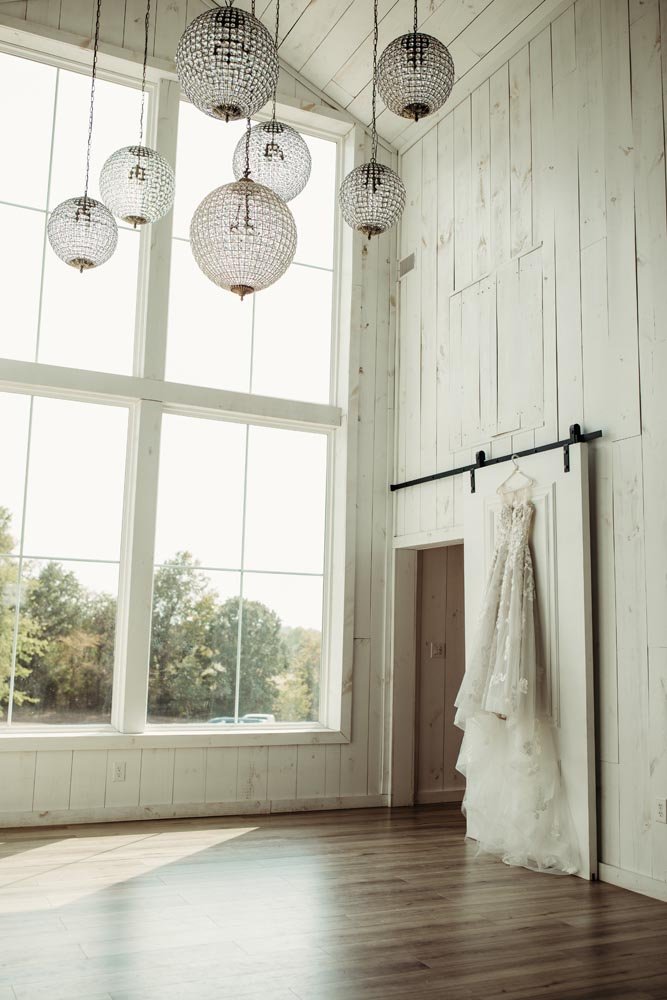 A white wedding dress hanging on a white wall on a sliding barn door, in a room with large windows and hanging spherical pendant lights.