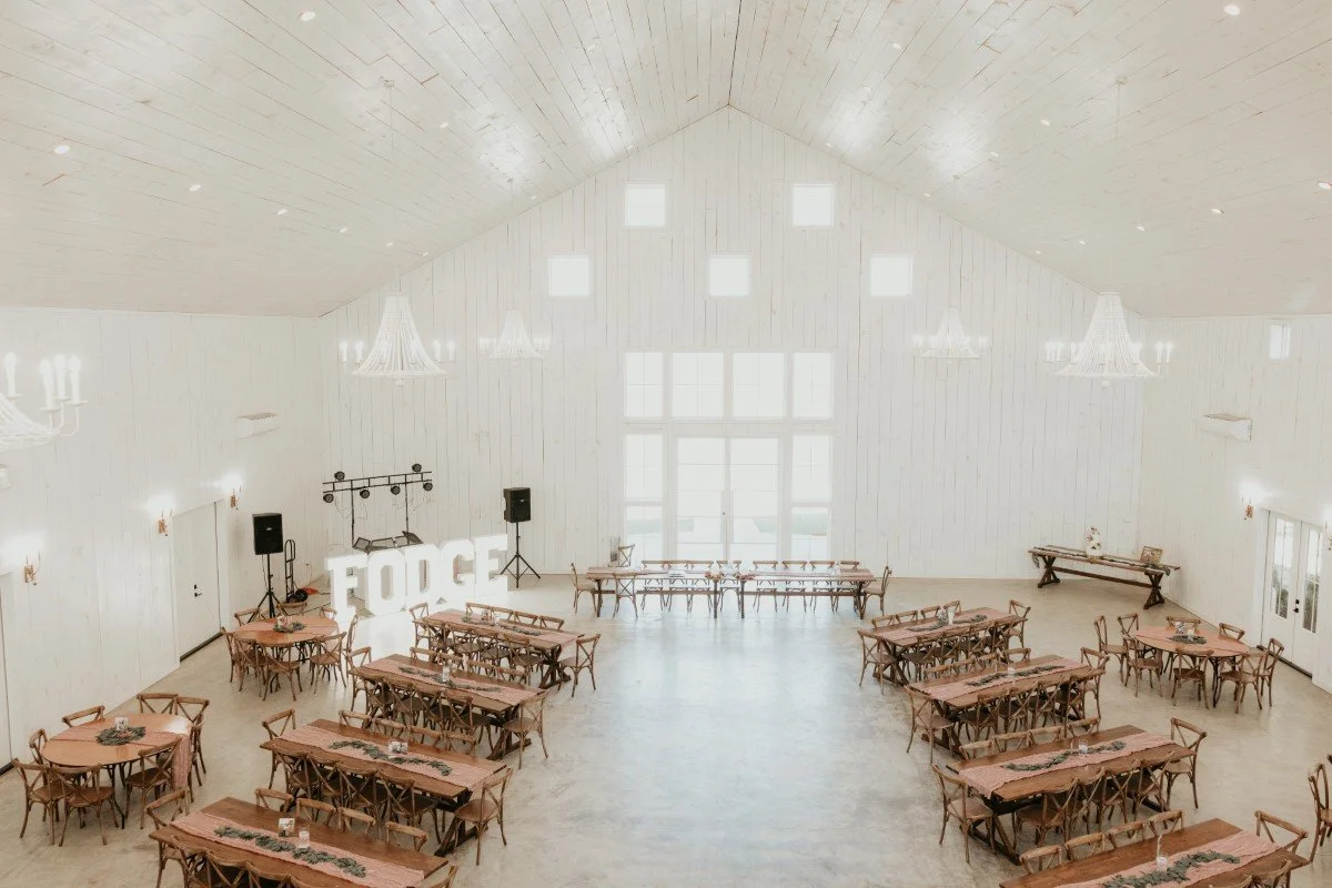 Large, bright event hall with white walls and ceiling, multiple chandeliers, tables with chairs arranged in rows, a DJ setup with speakers and large 'FORGE' sign, and a large window letting in natural light.