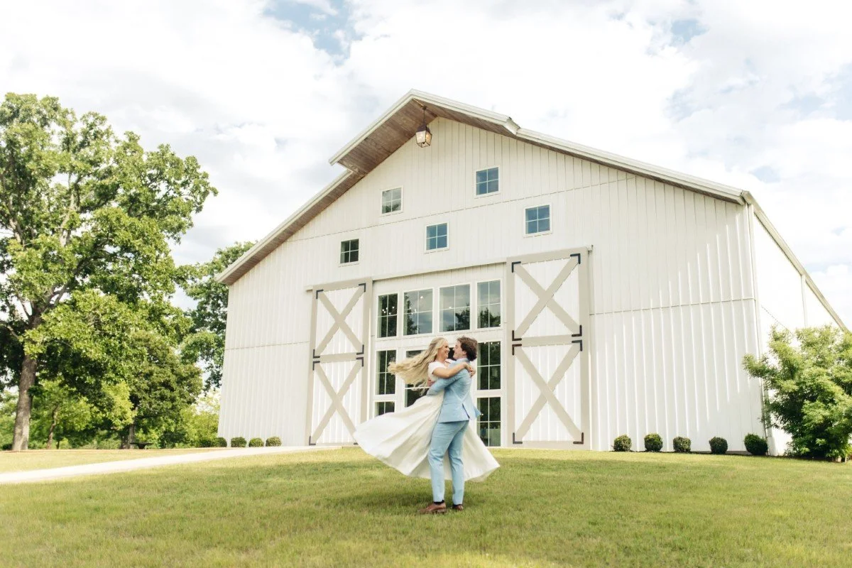 A couple dressed in wedding attire dancing in front of a large white barn with sliding barn doors, surrounded by trees and greenery.