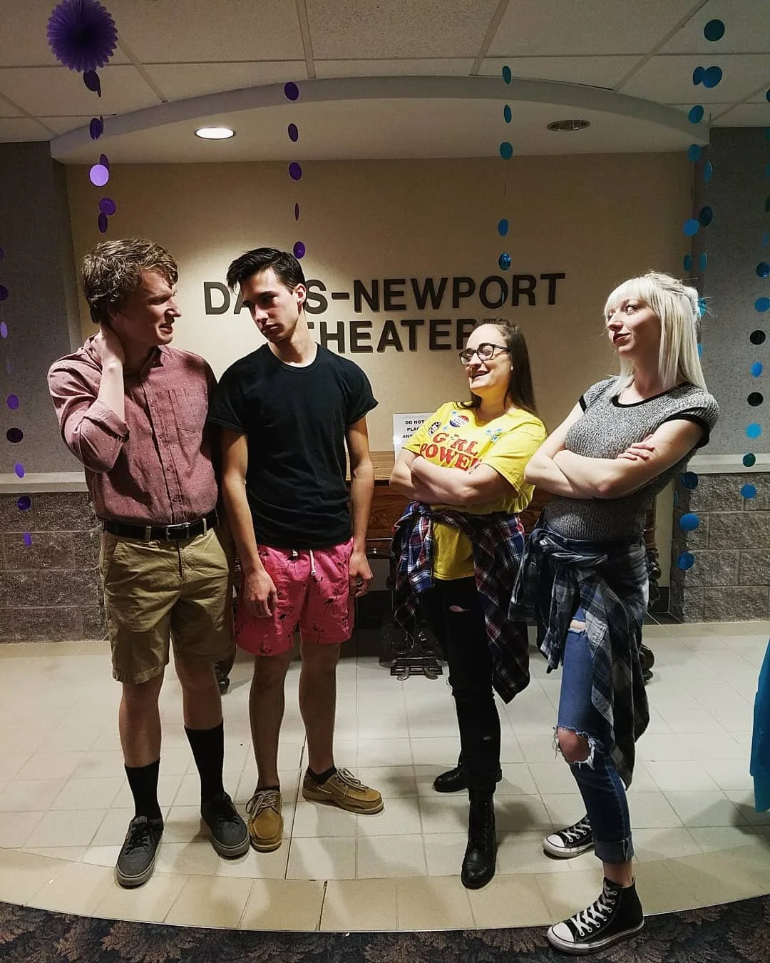 Four young people standing in front of a sign that reads 'DAYS-NEWPORT THEATER', with purple and blue decorations hanging from the ceiling.
