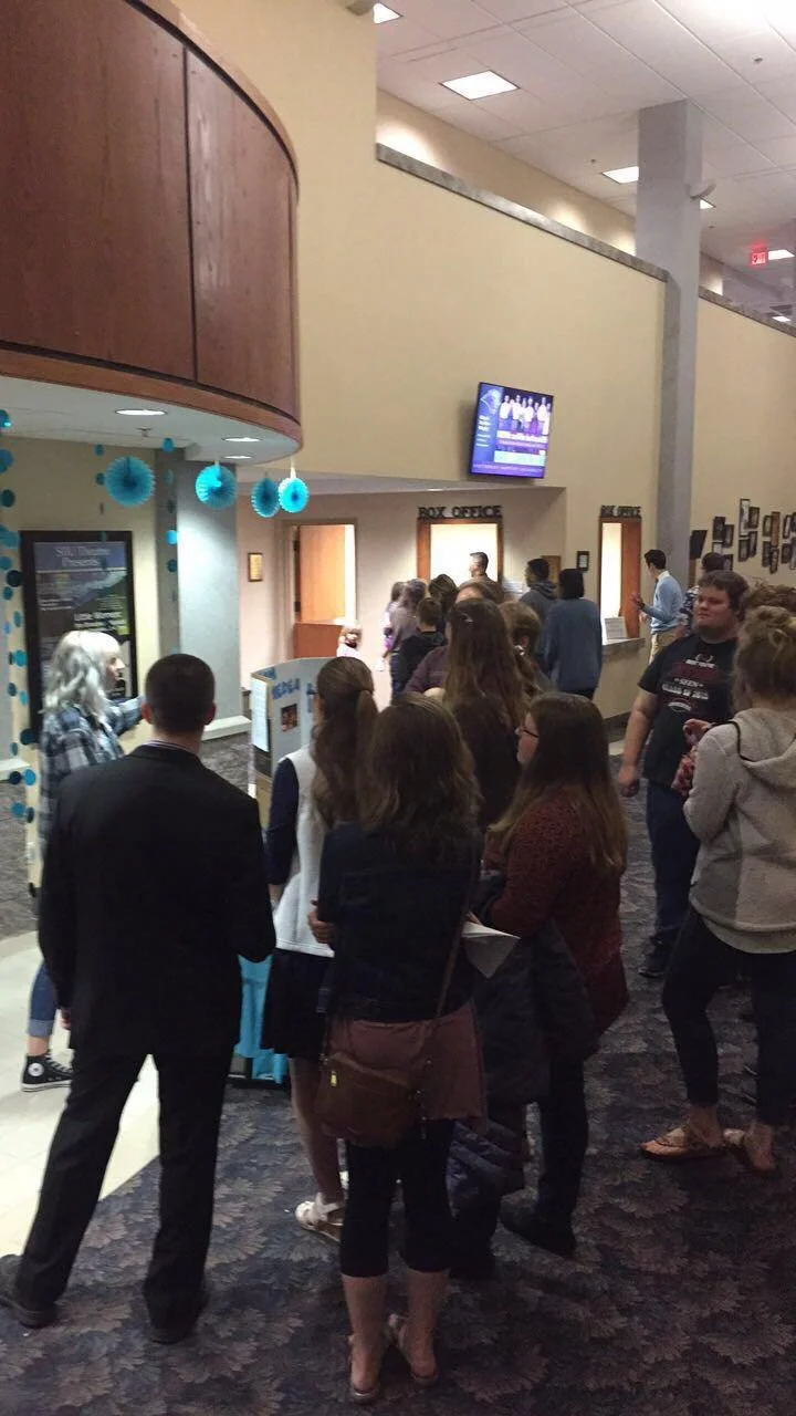 People standing in line at a box office inside a theater or event venue, with a decorated wall and a television screen above.