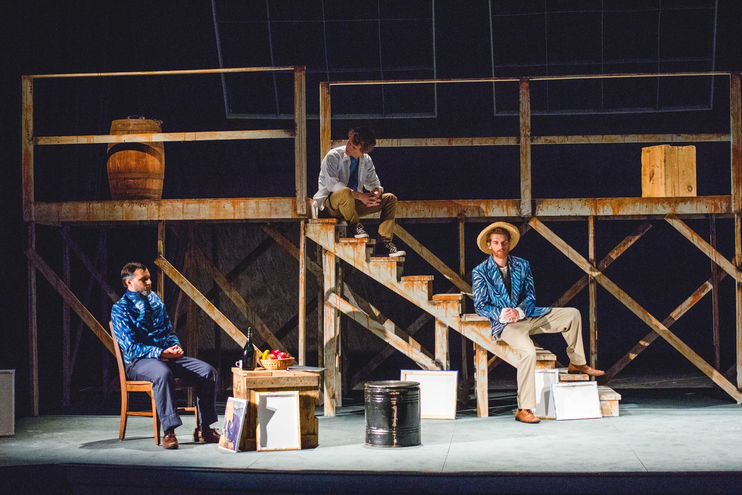 Four men on a stage set with wooden stairs and balcony, with props including a chair, table, barrel, bottles, fruit, and paintings, during a theatrical performance.