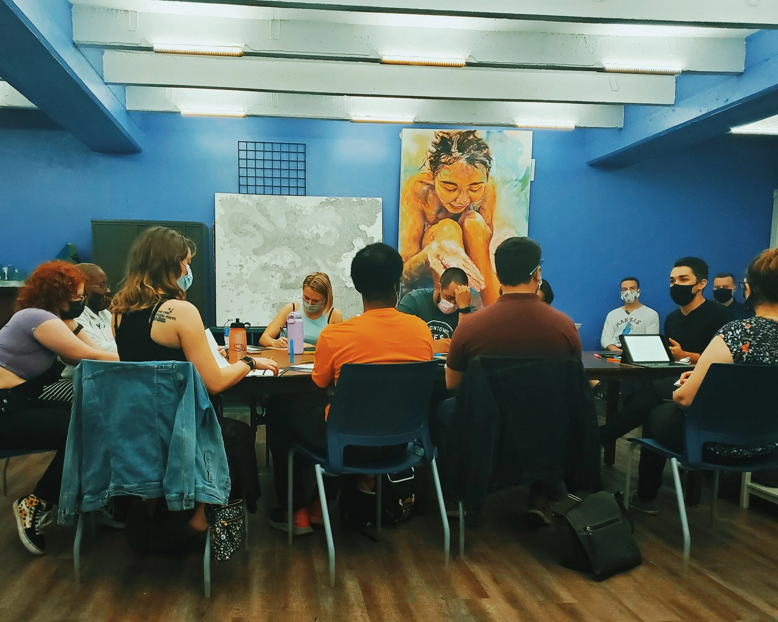 A rehearsal for Oedipus the King backstage at the Arts Asylum. The cast is gathered around the table for a reading. The cast are wearing COVID masks.