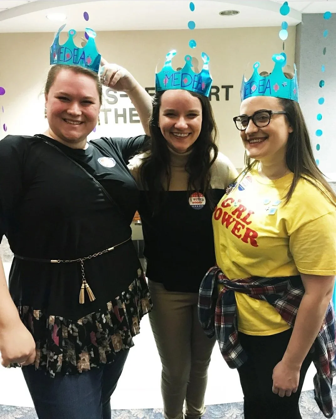 Three women wearing blue paper crowns with 'Team MEDA!' written on them, smiling and standing together at an event.