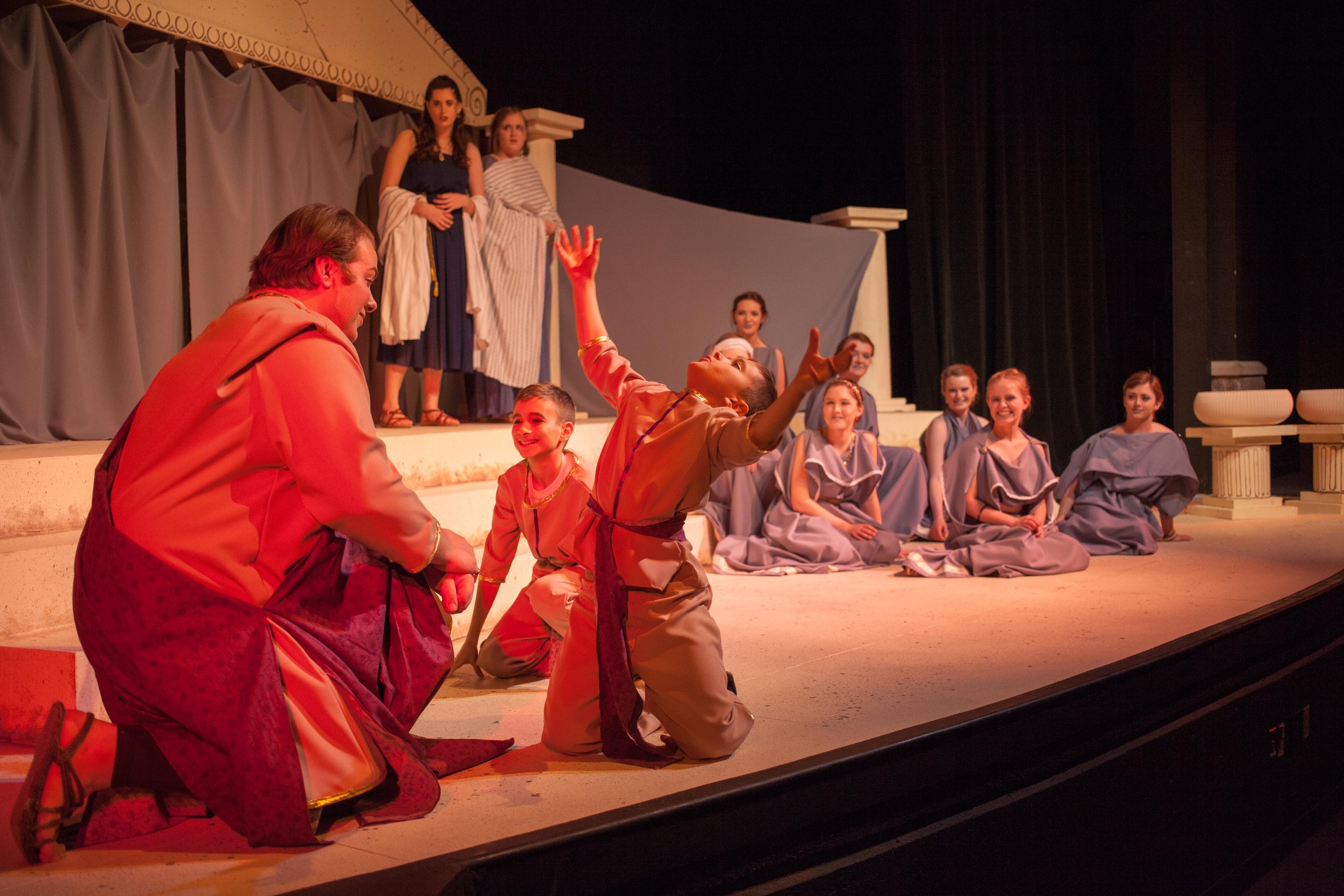 Children acting in a theatrical play on stage, with some kneeling and others standing, dressed in costumes, with a backdrop resembling ancient Greek or Roman architecture.