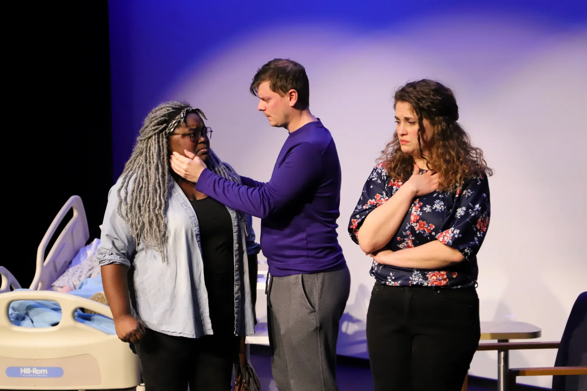 Three women and a man on stage during a theatrical performance. One woman is sitting in a hospital bed. The woman with gray dreadlocks and glasses appears distressed as the man gently holds her face. The third woman, with curly hair and a floral shirt, stands nearby with a concerned expression.