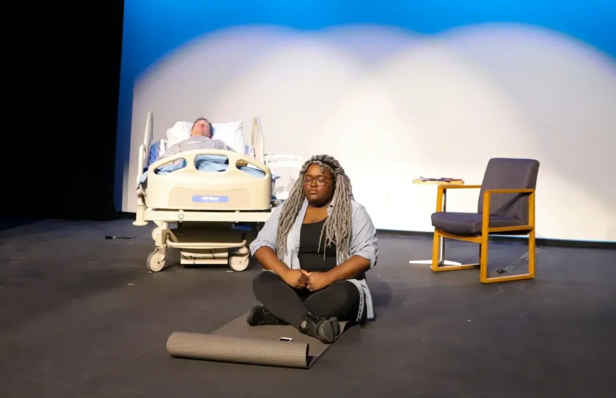 In the foreground, an actress is doing yoga on a yoga mat. In the background, an actor is lying in a hospital bed. Production photo from KC Public Theatre's production of The Holy Trinity with dramaturgy by Soph Marsh