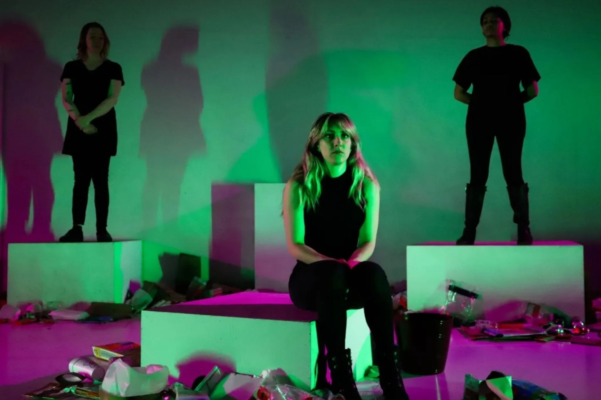 A production photo from MIRRORS by Kaitlin Gould at KC Public Theatre. A blonde woman sits on a cube center stage, surrounded by trash. Behind her, two actors dressed in all black loom ominously.