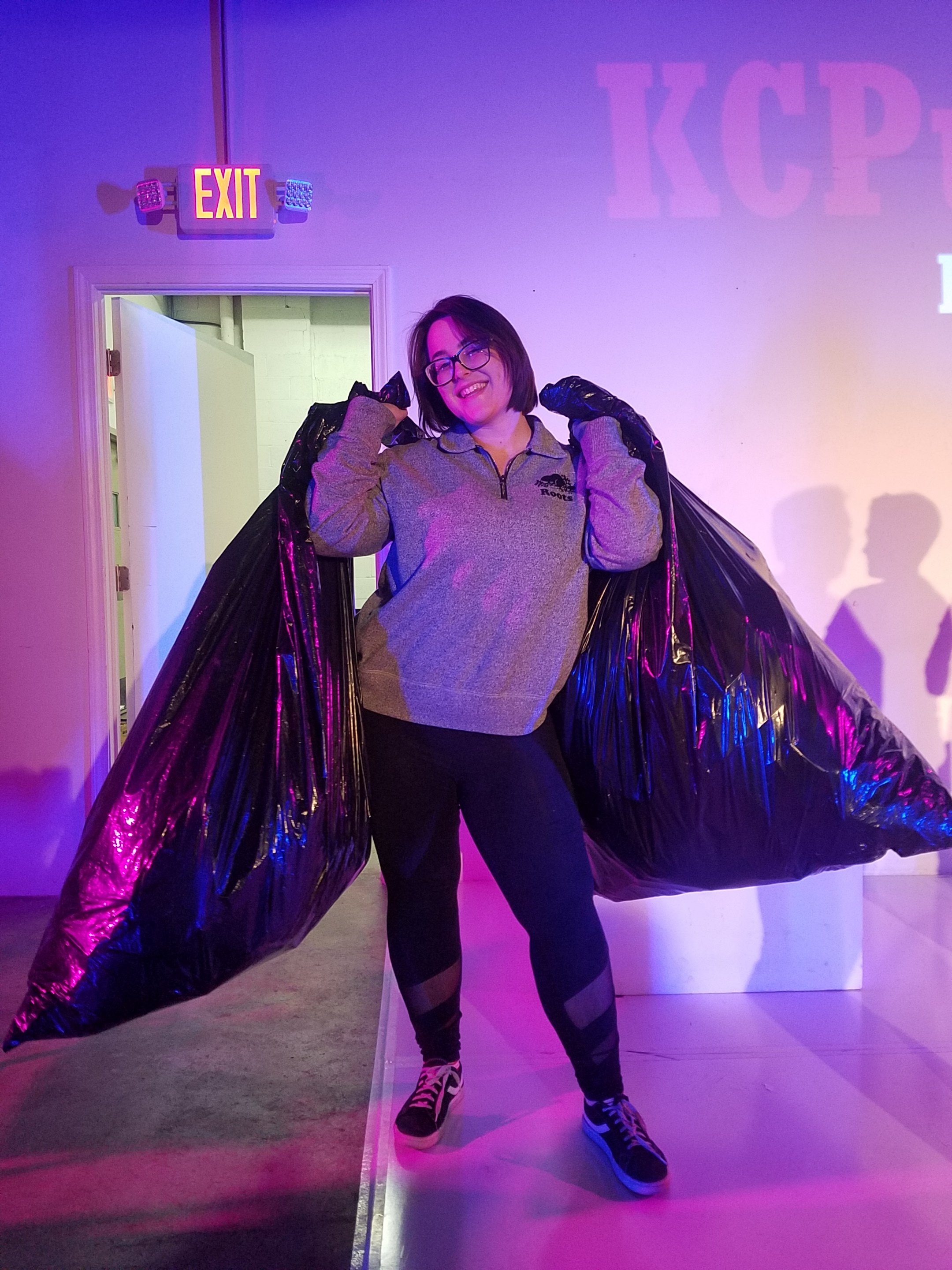 A smiling woman with glasses carrying two large black garbage bags, standing indoors with colorful lighting and a sign above the doorway.