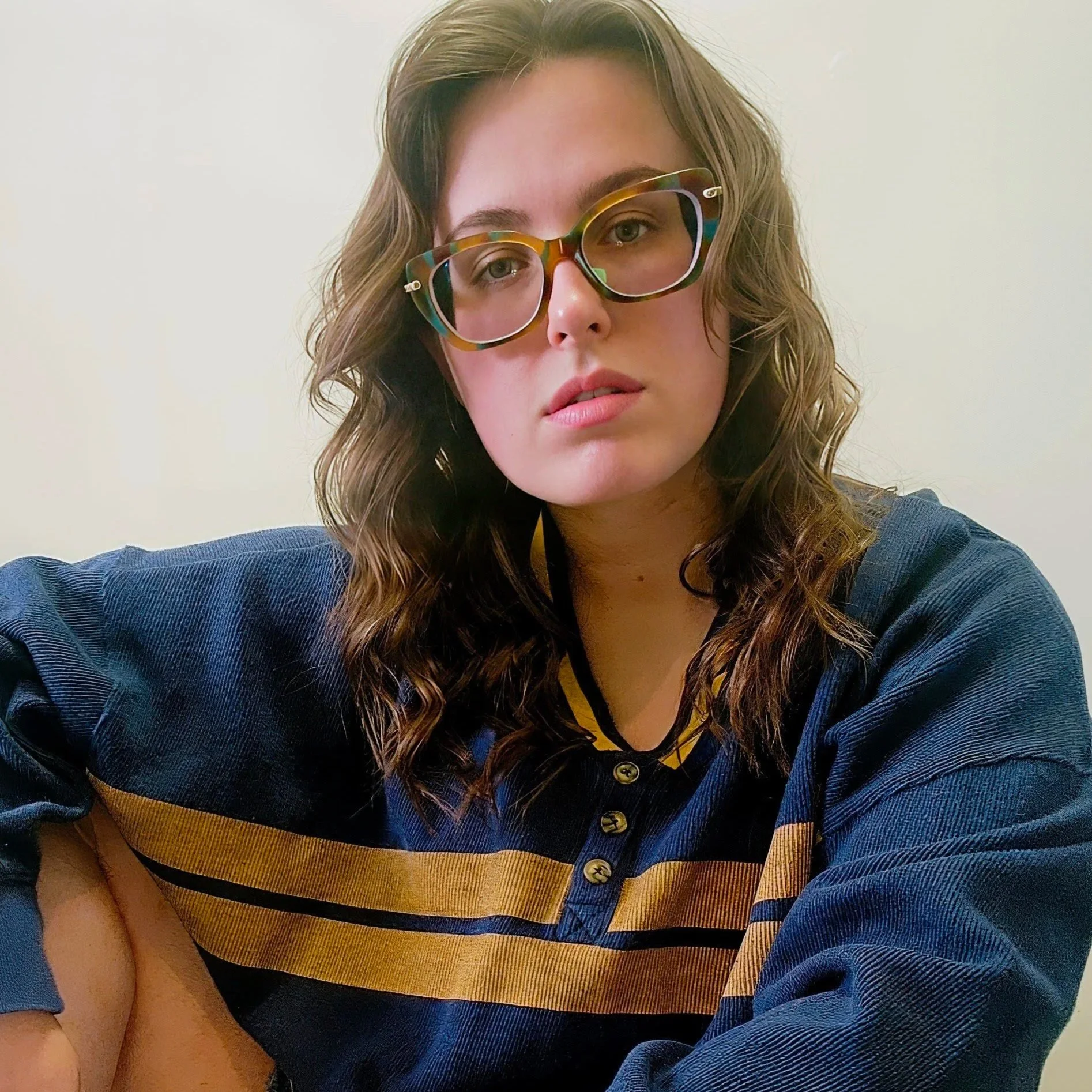 soph marsh's headshot. They have curly brown hair, turtle-rimmed glasses, blue eyes. They're pictured wearing a blue and yellow striped sweater.