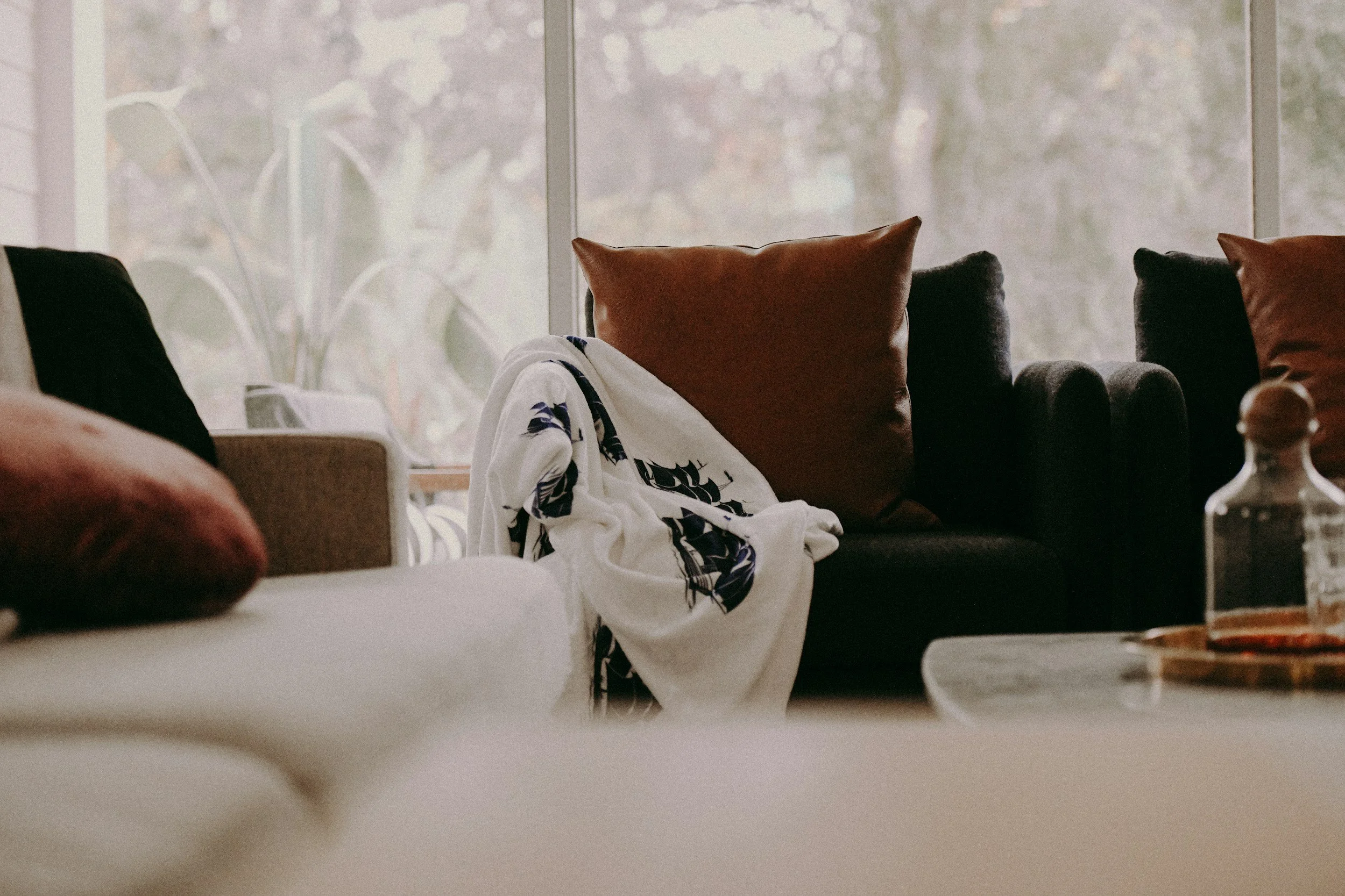 A cozy living room with a black sofa, brown pillows, a white blanket, and a glass jar on a coffee table in front of a large window showing trees outside.