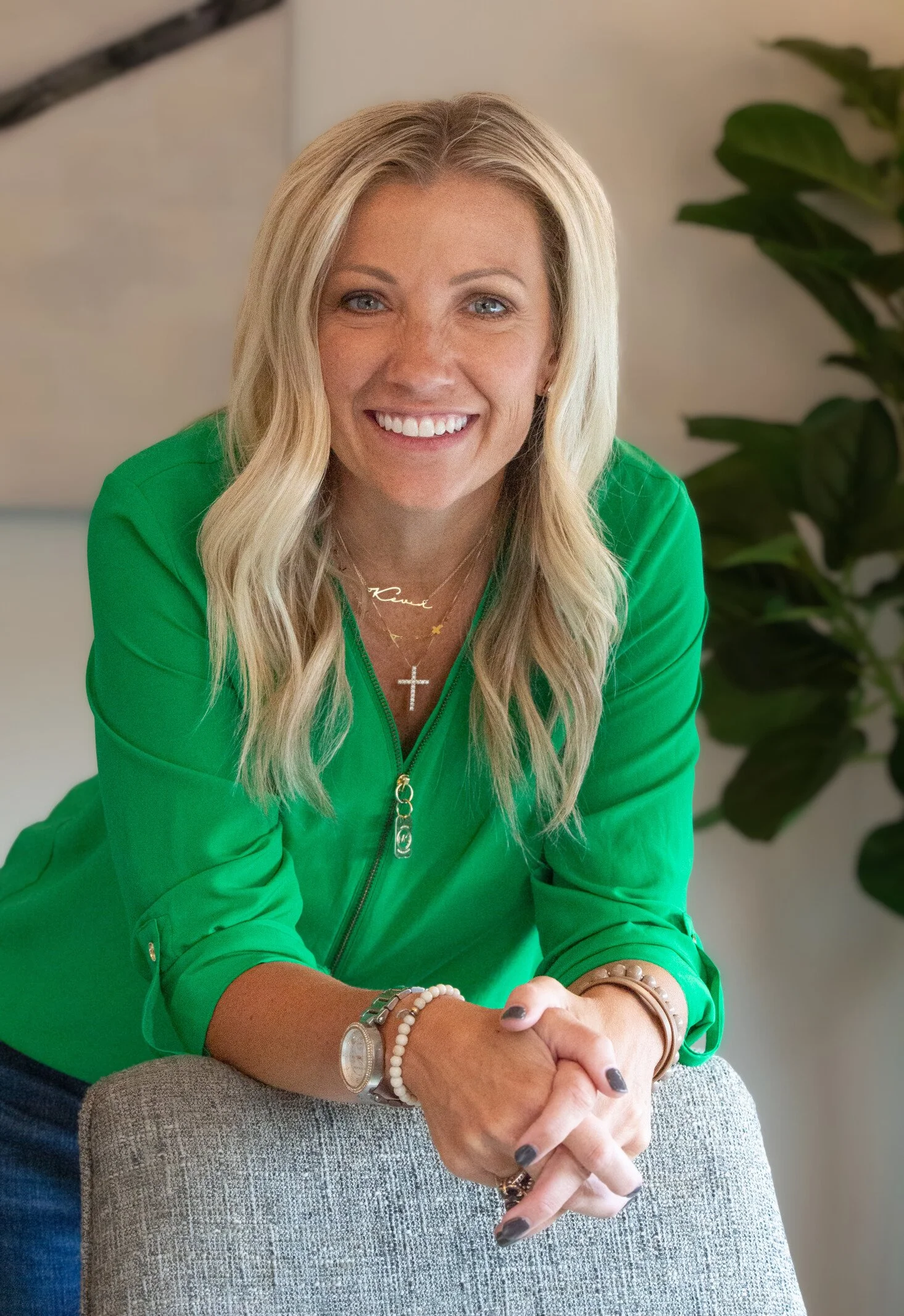 A woman with blonde hair in a green jacket smiling and sitting with her hands clasped on a chair armrest, in front of a houseplant.