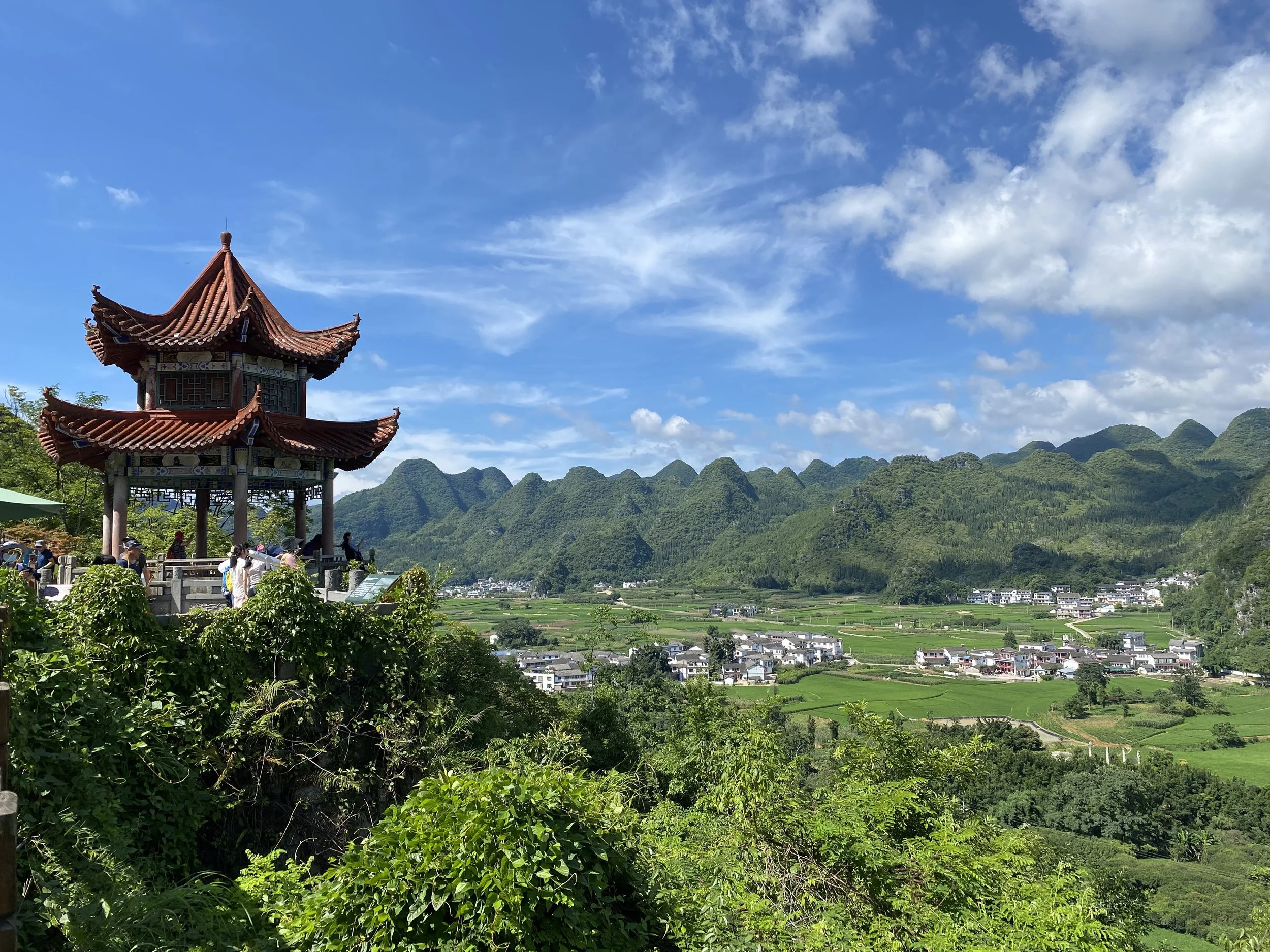 A traditional Chinese-style pavilion with a pagoda roof on a hill overlooking green farmland and distant mountains under a cloudy blue sky in Guizhou province, China