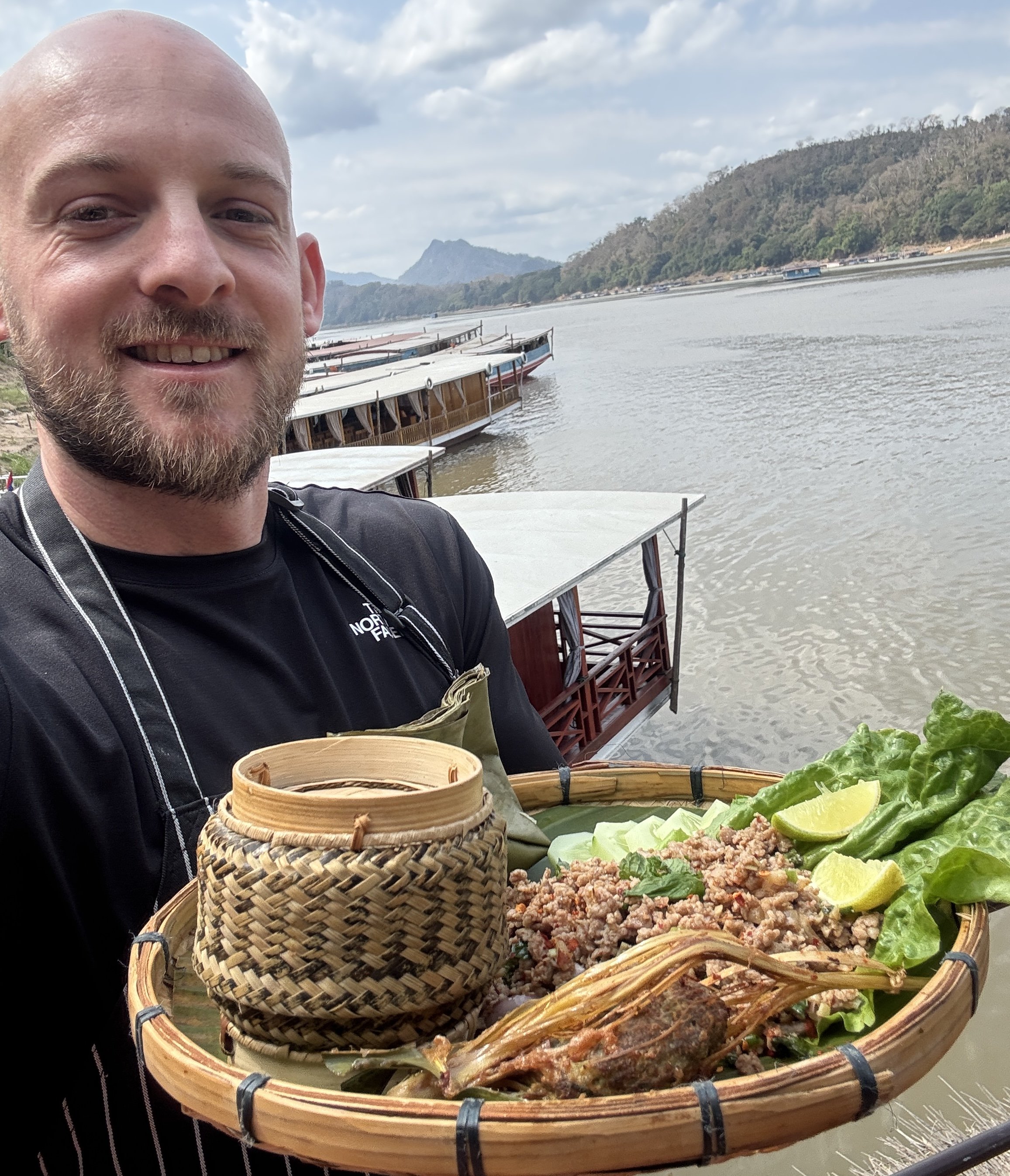 A smiling man holding a woven tray of fresh Laotian larb salad, herbs, and a bamboo sticky rice basket on a Mekong river boat.