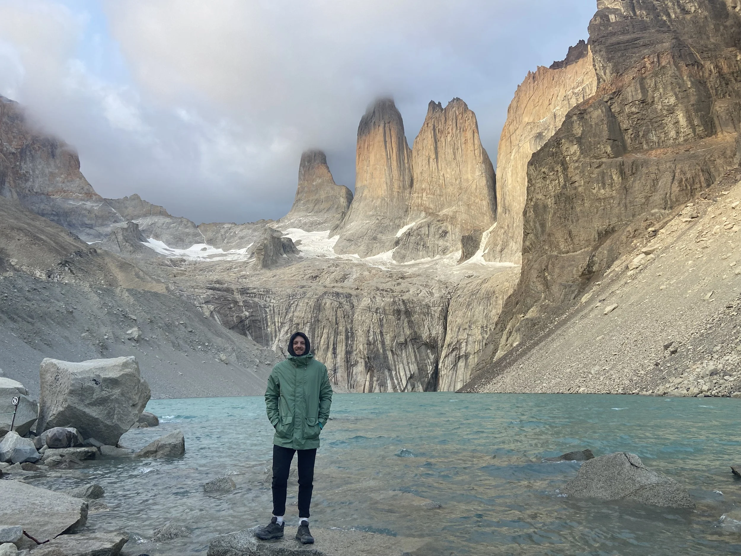 Holocene founder standing in front of a lake with mountains of Torres Del Paine in Chile in the background. The person is wearing a green jacket and black pants, and the sky is cloudy.
