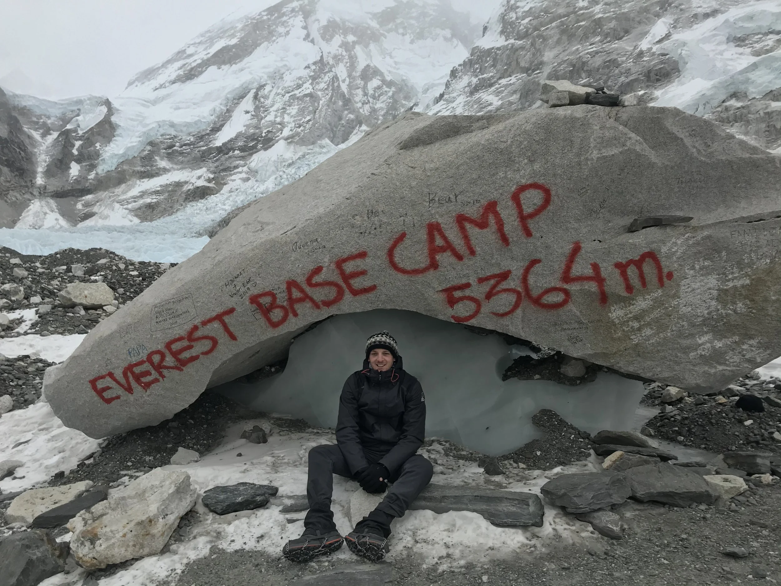 Holocene founder sitting in front of a large boulder that reads 'EVEREST BASE CAMP 5364m' at a mountain glacier after trekking in the Himalayas of Nepal, with snow-covered peaks in the background.