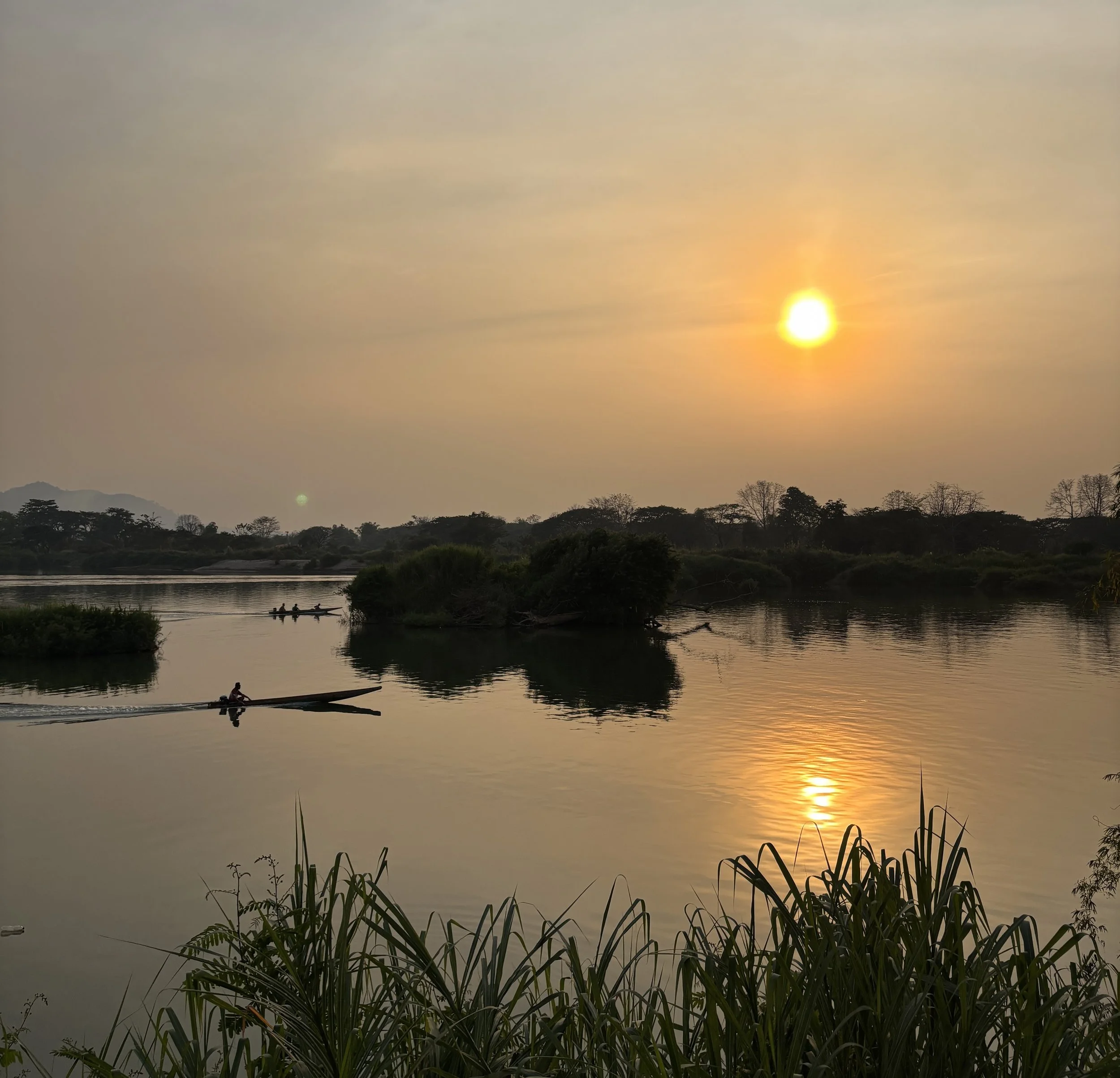 A brilliant golden sunset reflecting on a wide river as longtail boats cruise past small, lush islands in Laos.