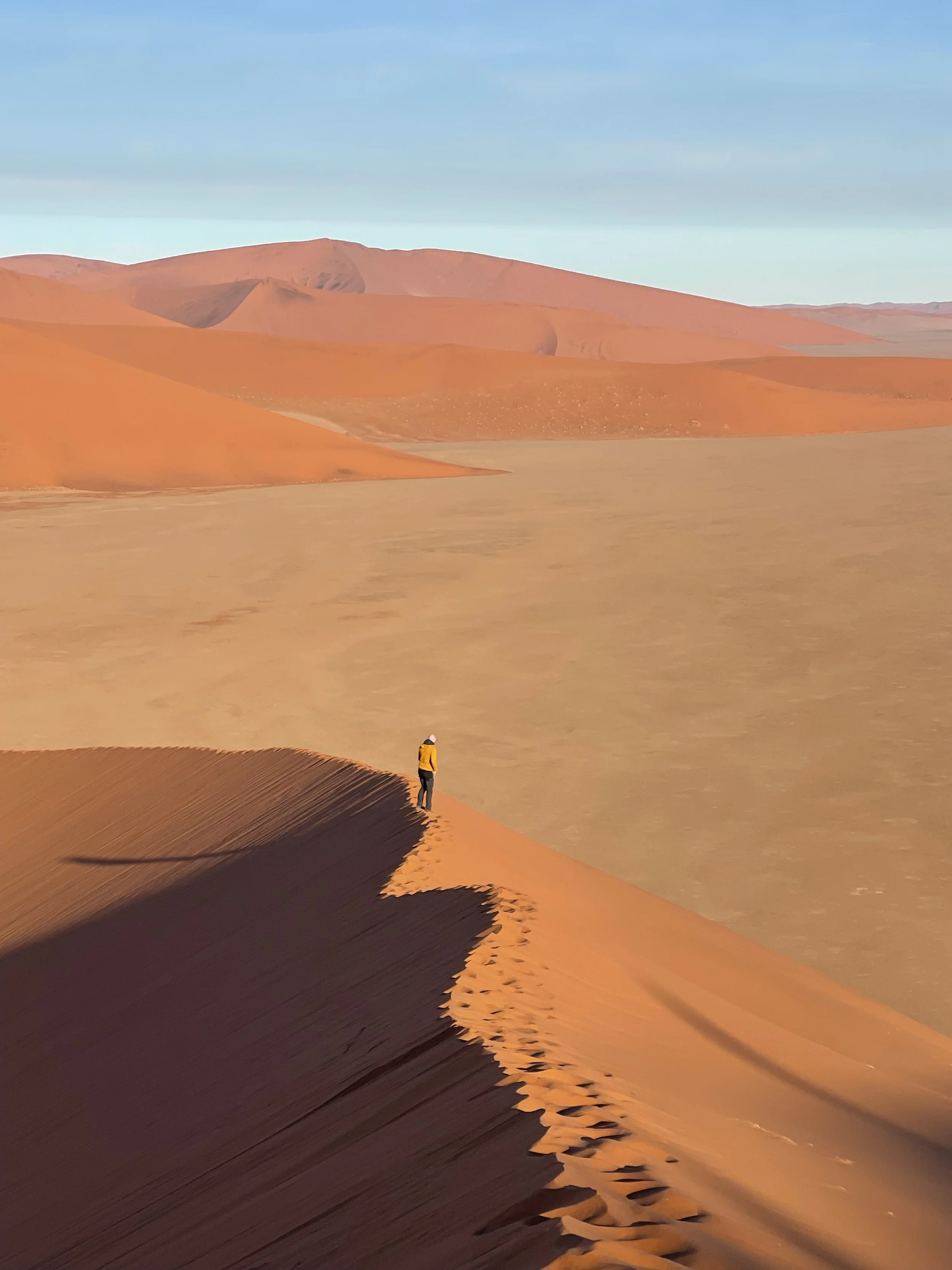 Holocene founder in a yellow jacket standing on a sand dune in a desert in Namibia with rolling sand dunes extending into the distance and a blue sky above.