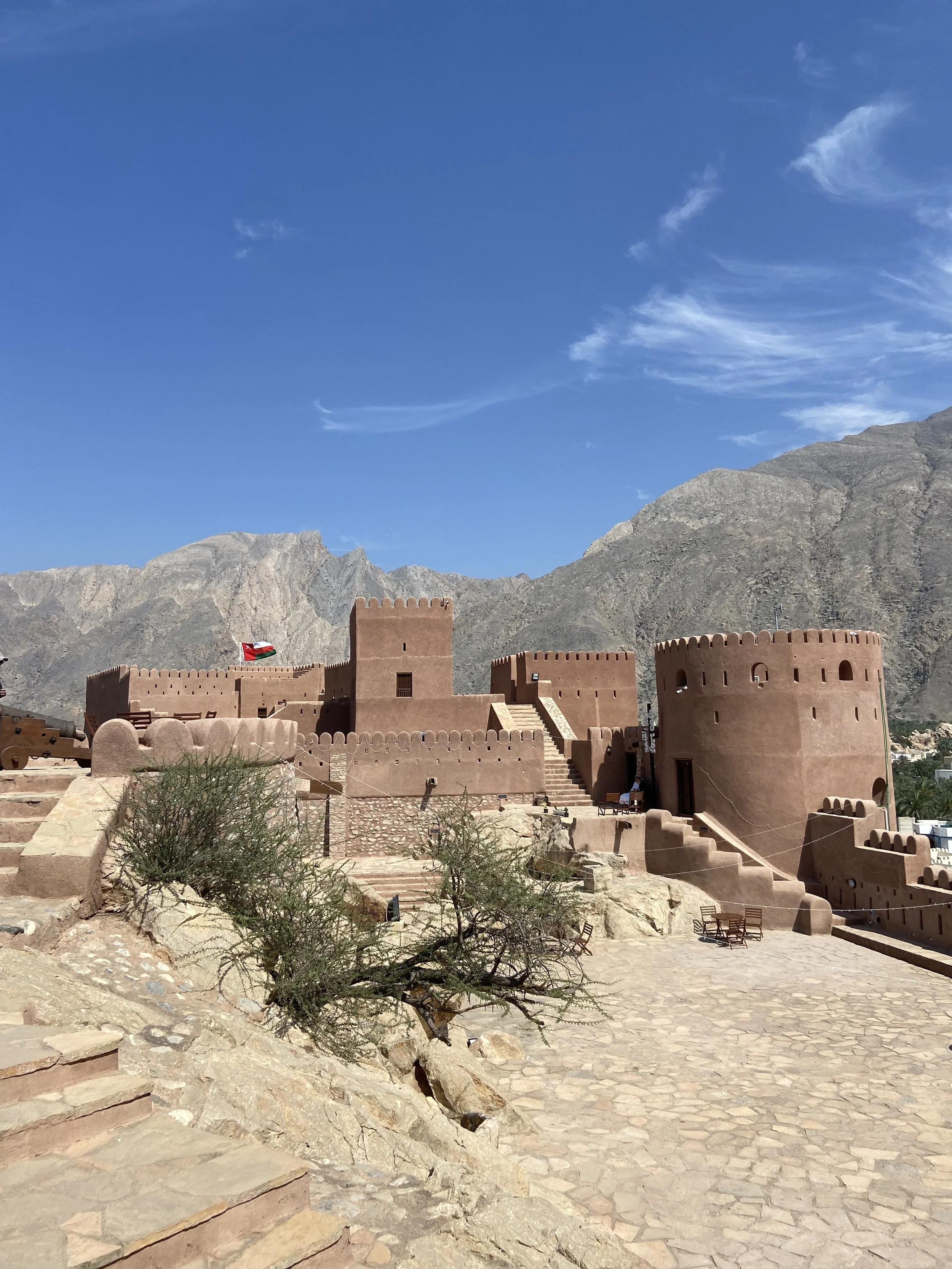 An ancient stone fortress with rounded towers, a flag flying from one tower, set against a backdrop of rugged mountains and a clear blue sky with wispy clouds in Oman.