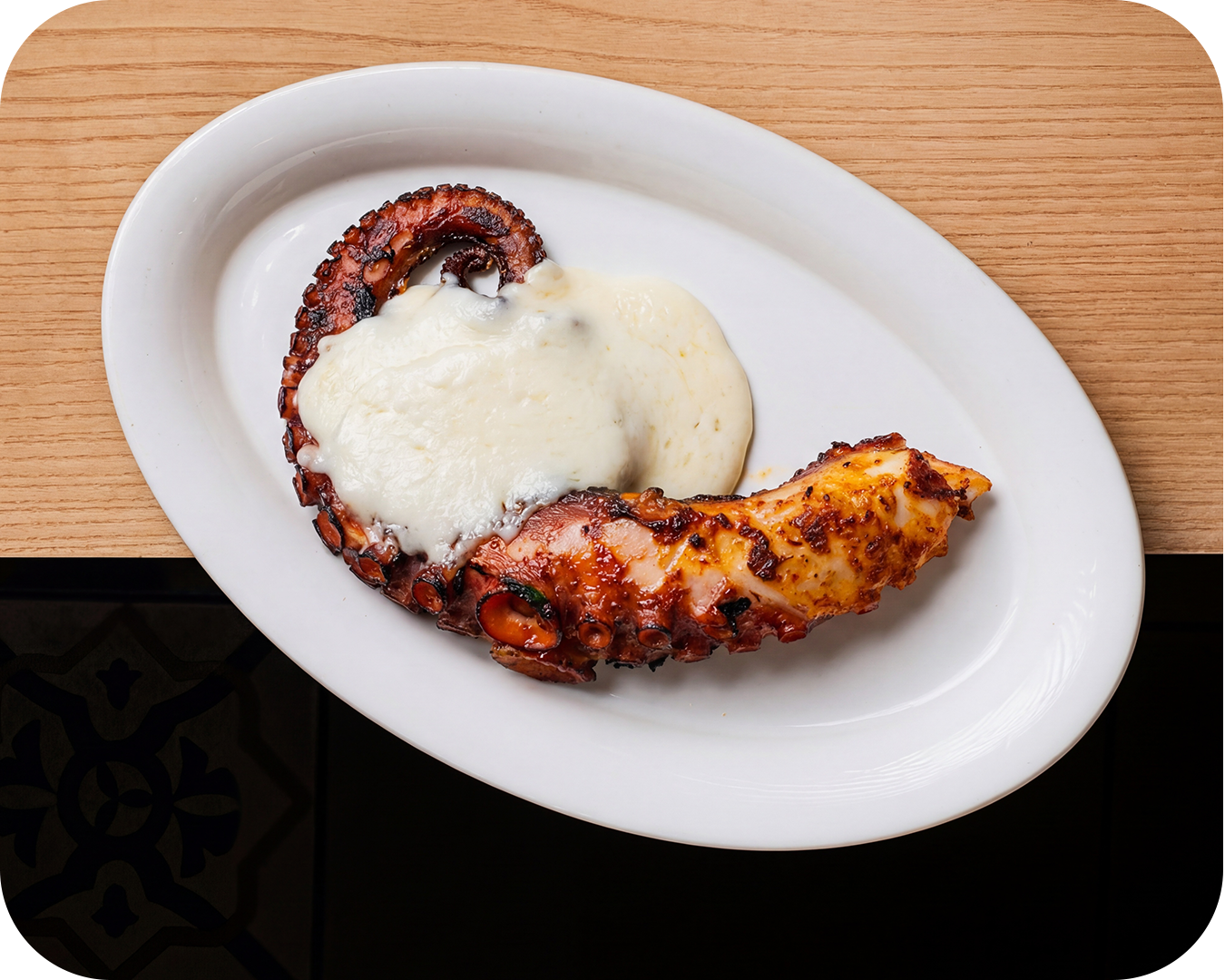 Pulpo a la parrilla cubierto con queso, en plato blanco sobre mesa de madera.