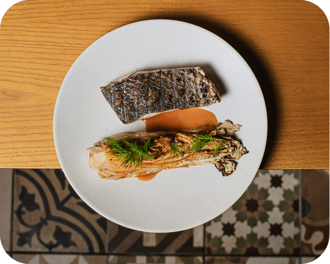 Pescado con piel crujiente y pescado en salsa con eneldo decorativo en plato blanco sobre mesa de madera.