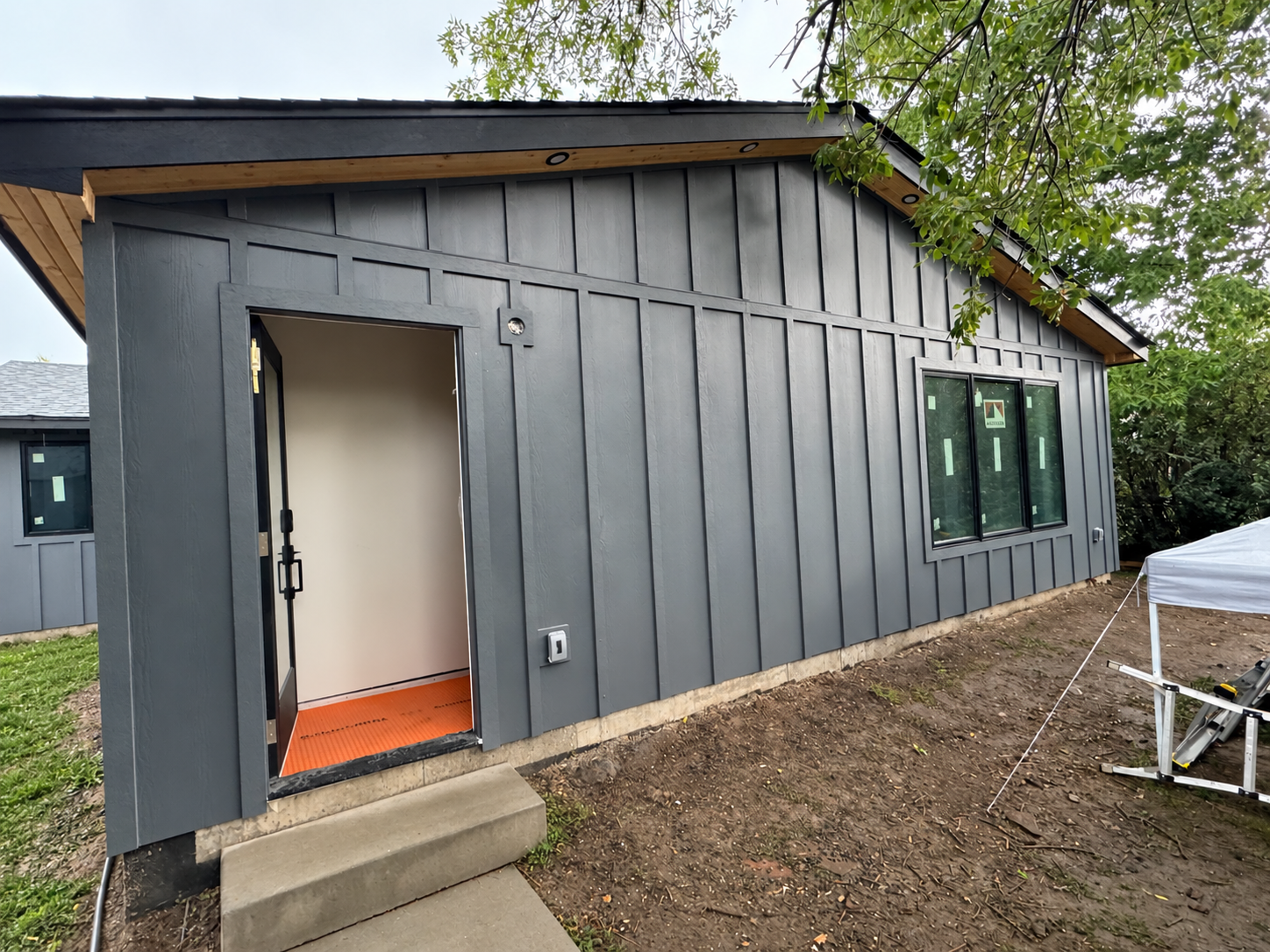 A house under construction with dark grey vertical siding, a window, and a front door with an orange mat, surrounded by trees and part of a portable work table.