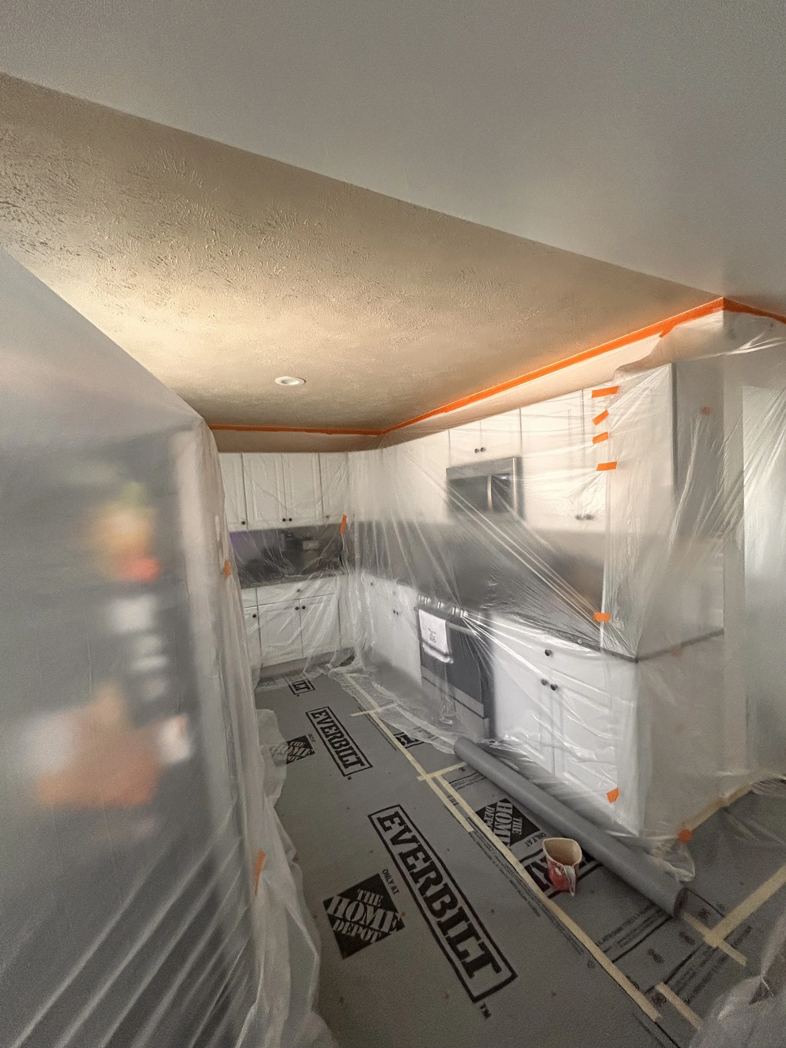 Interior view of a room under renovation with plastic sheeting covering the walls and cabinets, and construction materials and tape on the floor.