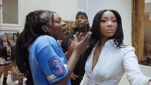 Two women having a conversation, one appears to be speaking passionately while the other listens with a neutral expression. A man is standing behind them, observing.