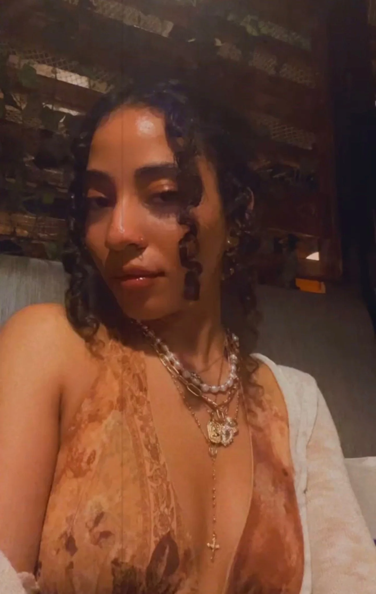 A woman with dark curly hair wearing layered necklaces and a sleeveless patterned top. She is sitting indoors against a wood-paneled background.